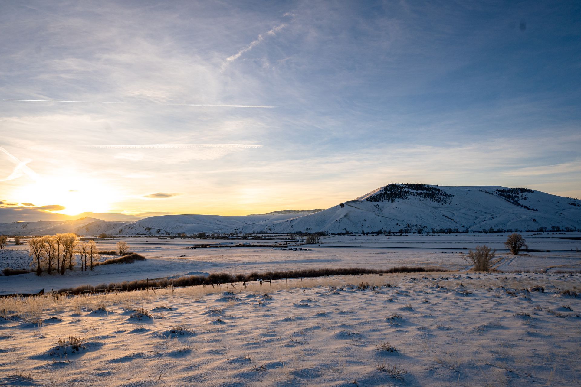 Snow-covered field at sunset with a hill in the distance. Pale blue sky, golden sunlight.