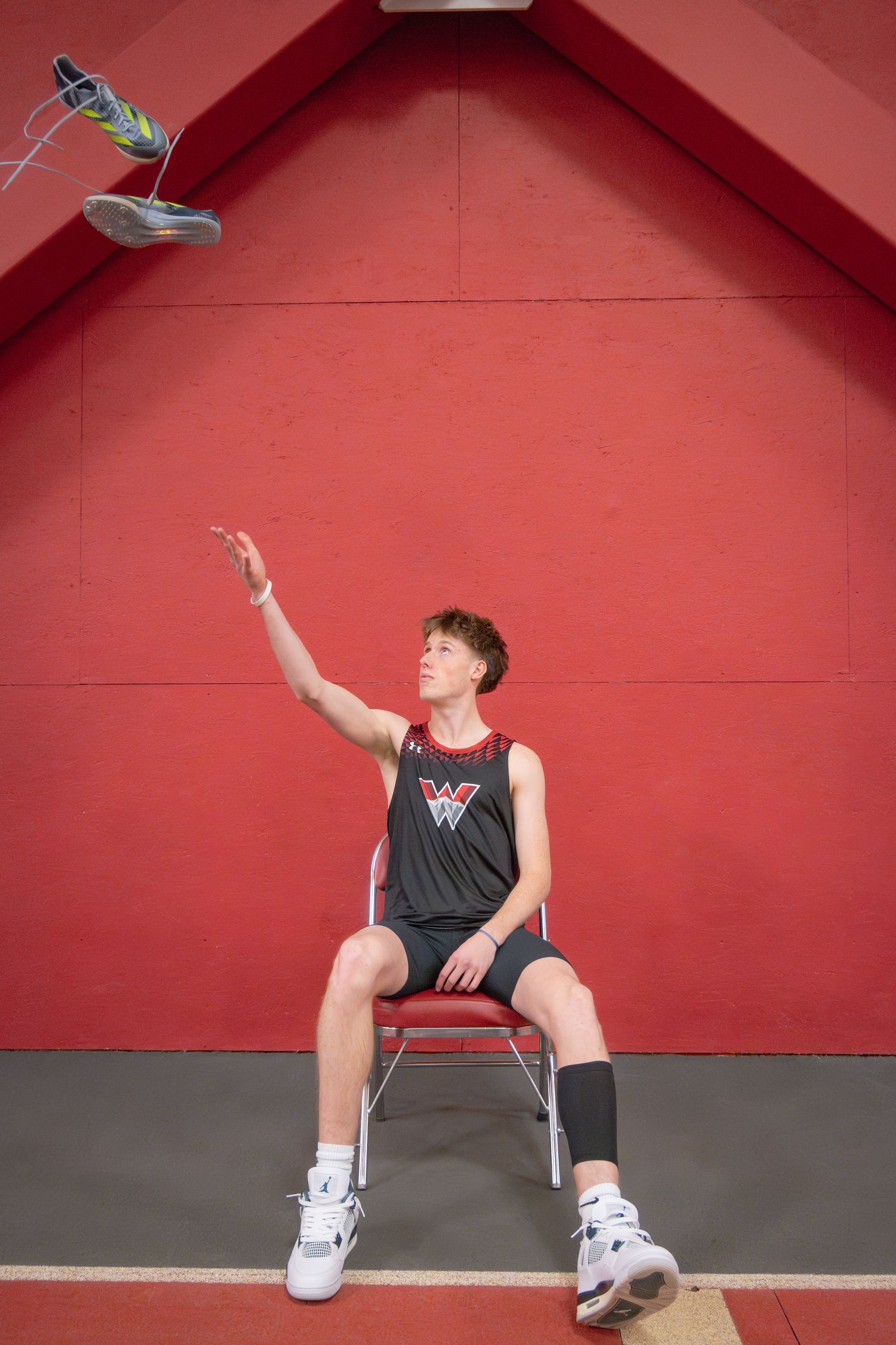 Teenager seated, throwing shoes in the air, against a red wall background.