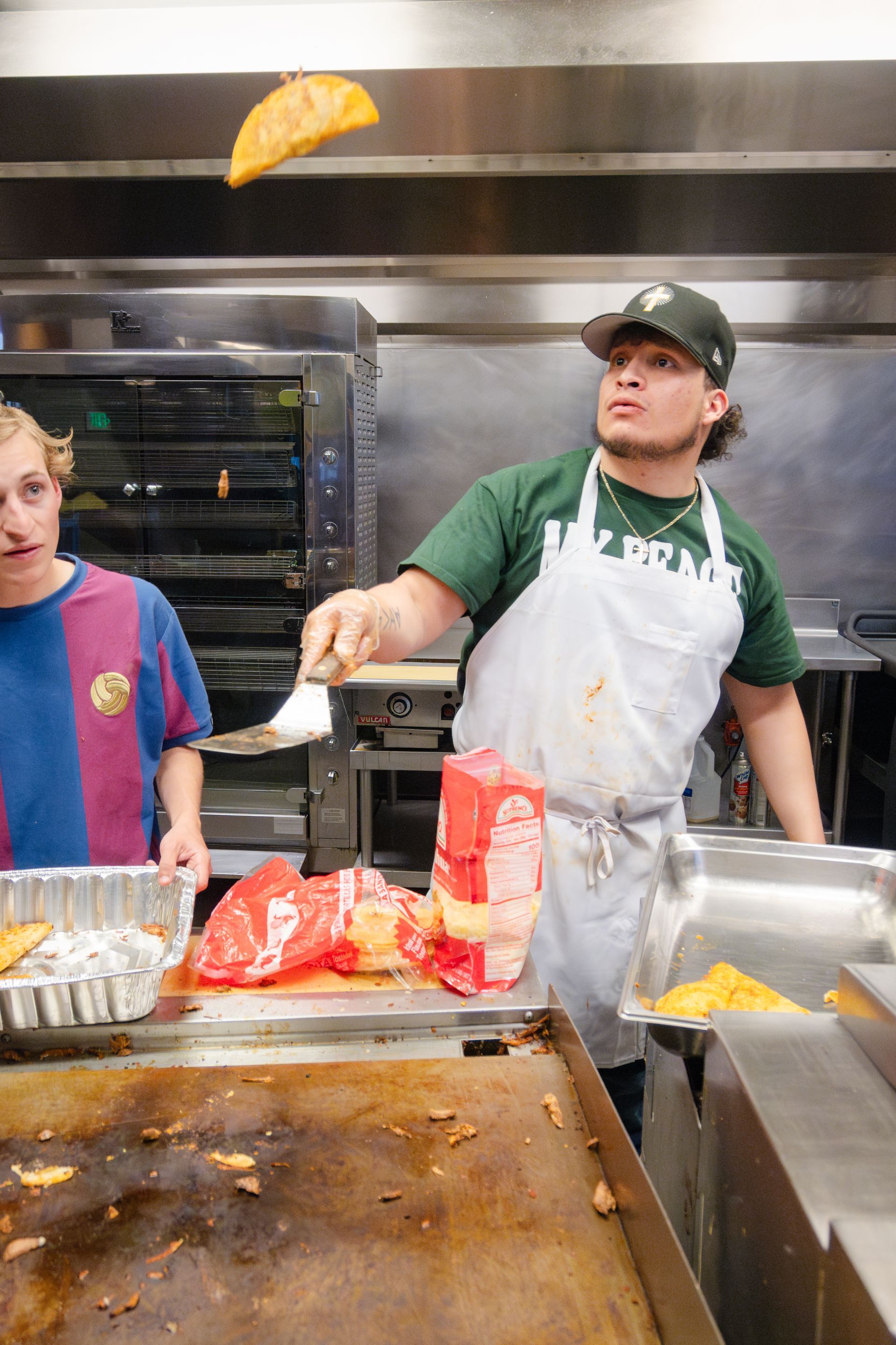 Chef tosses a taco in the air with a spatula. Kitchen setting, another person watches.