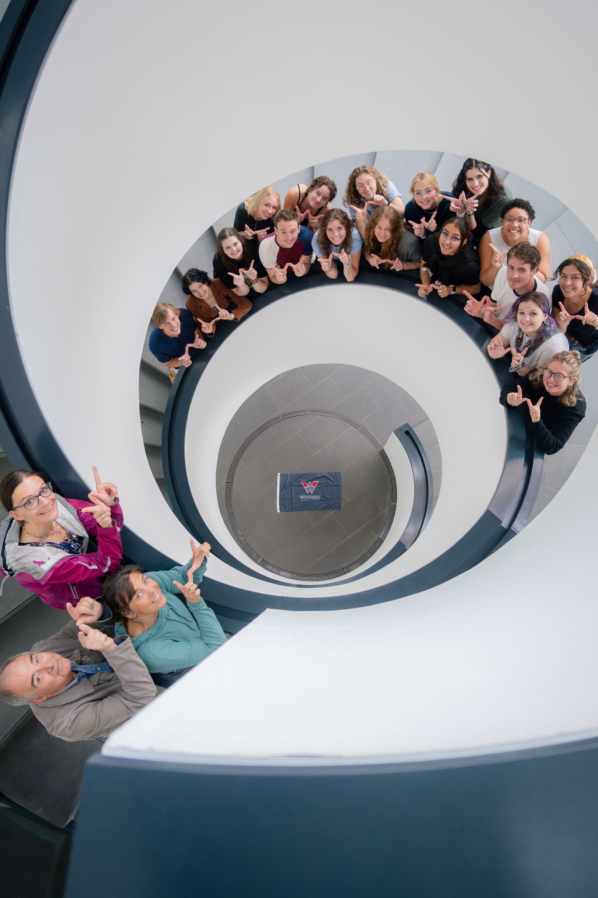 People looking down a spiral staircase, pointing towards center. Bright, white stairs, blue accents.