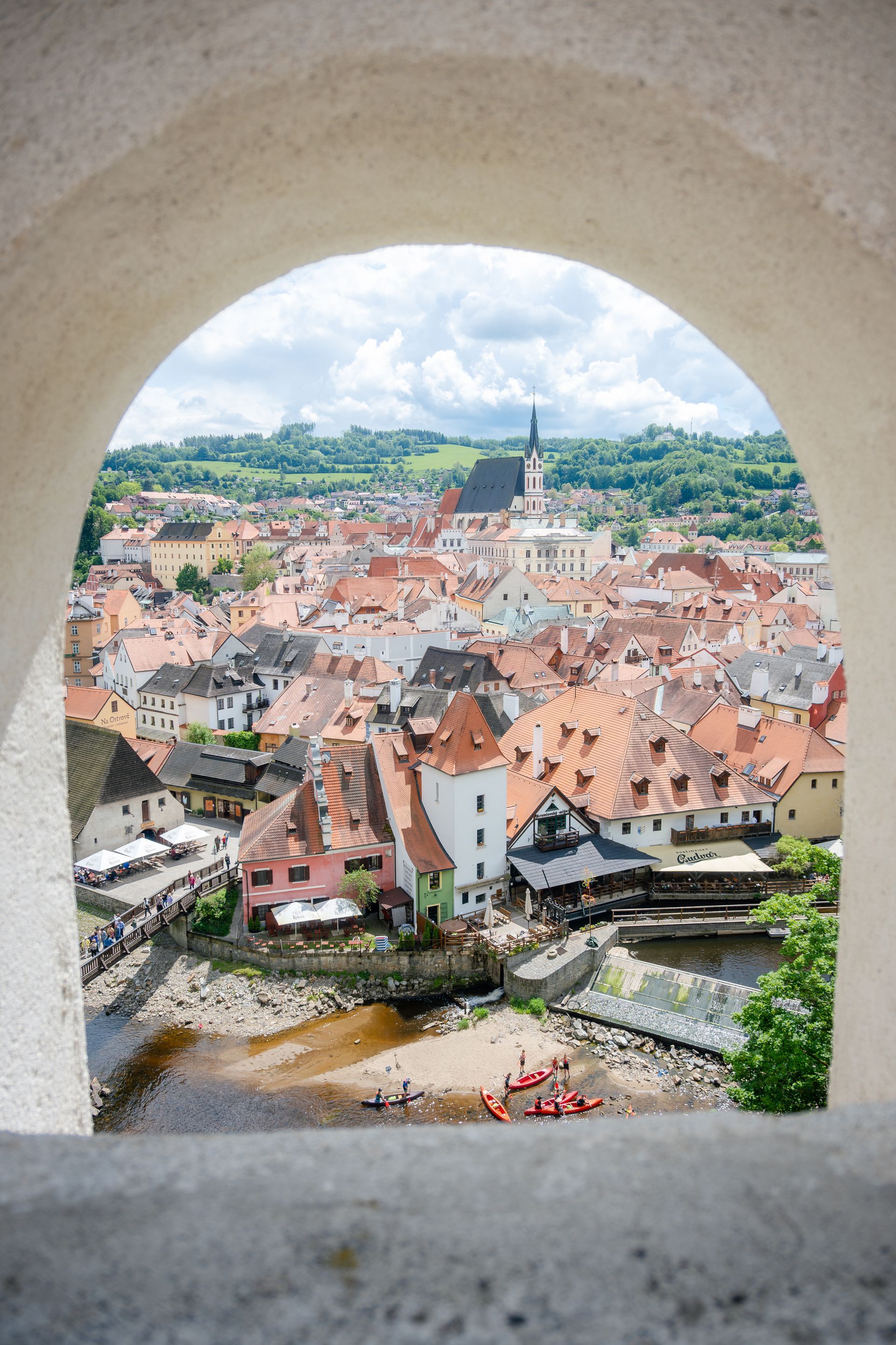 View of Cesky Krumlov from an arched window, red-roofed buildings, river, and green hills on a bright day.