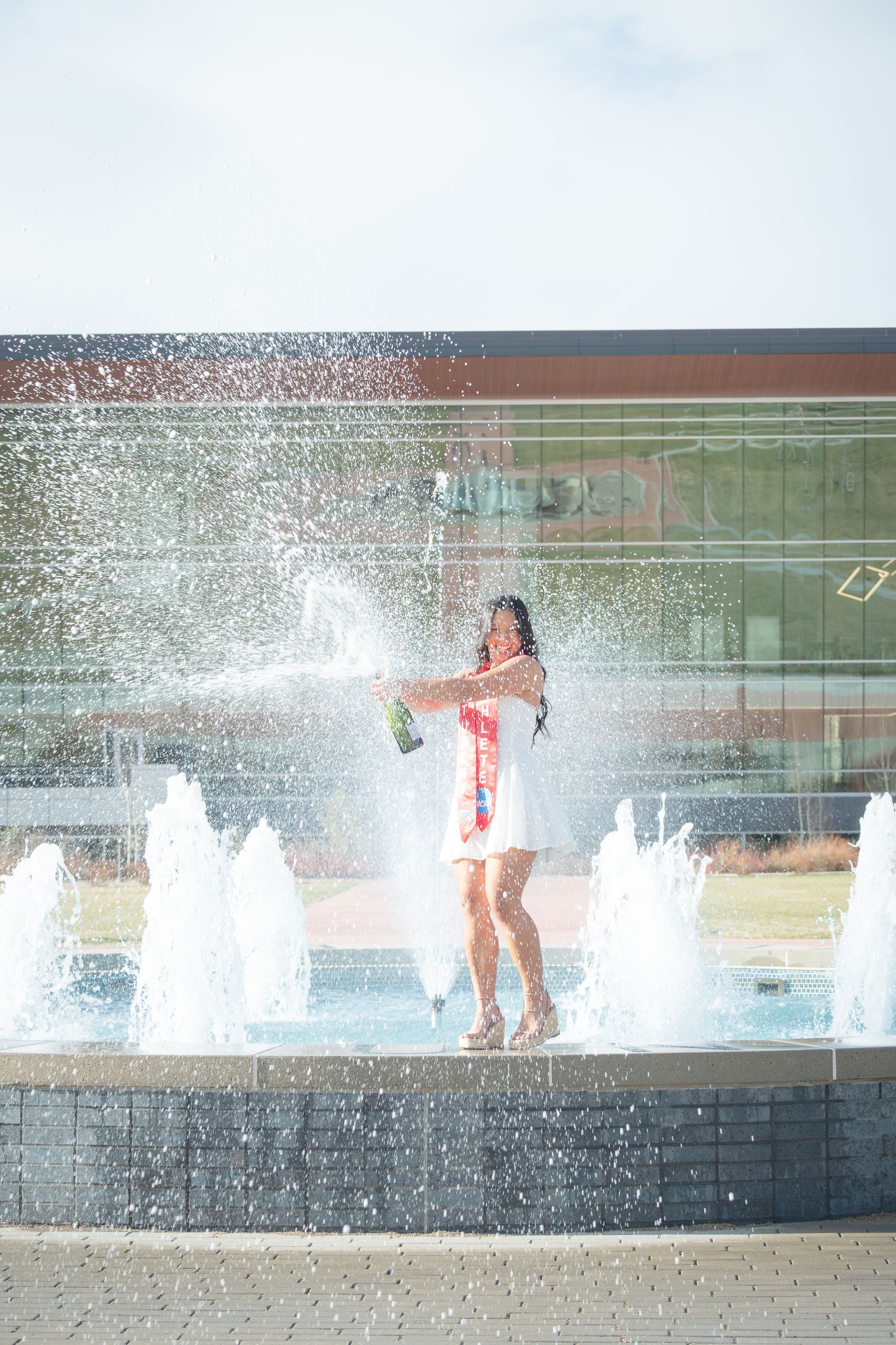 Woman in white dress pops champagne by a fountain on a sunny day, celebrating graduation.