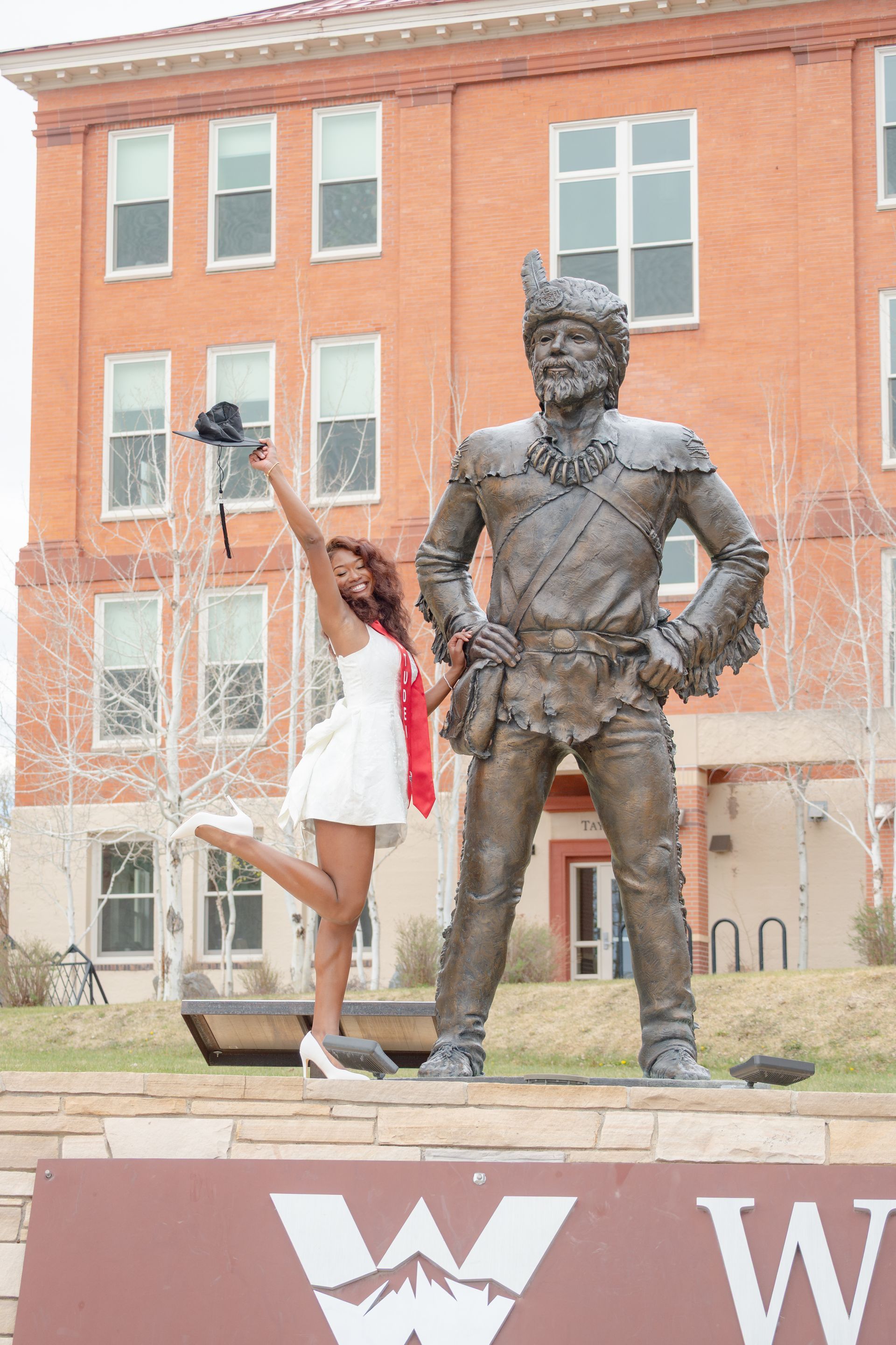 Woman in white dress poses with bronze statue outside a brick building.