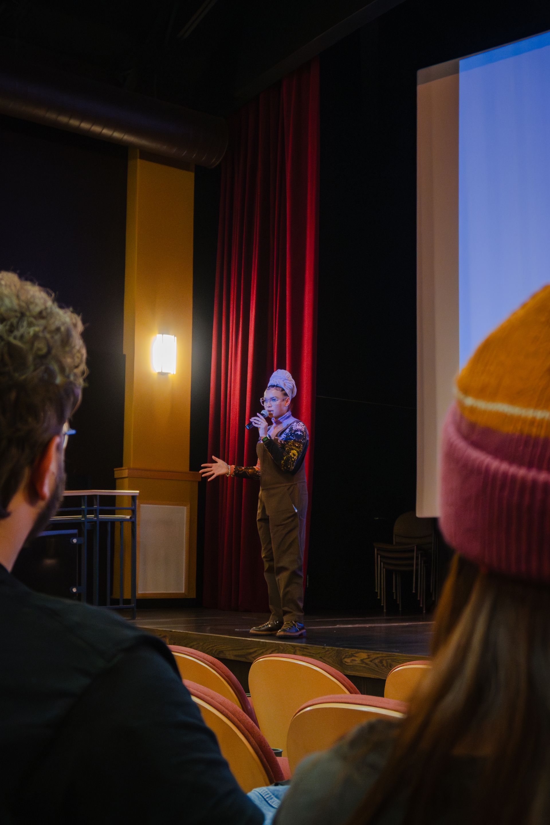 Person speaking on a stage, arms outstretched. Audience members in foreground. Stage has a screen and red curtains.