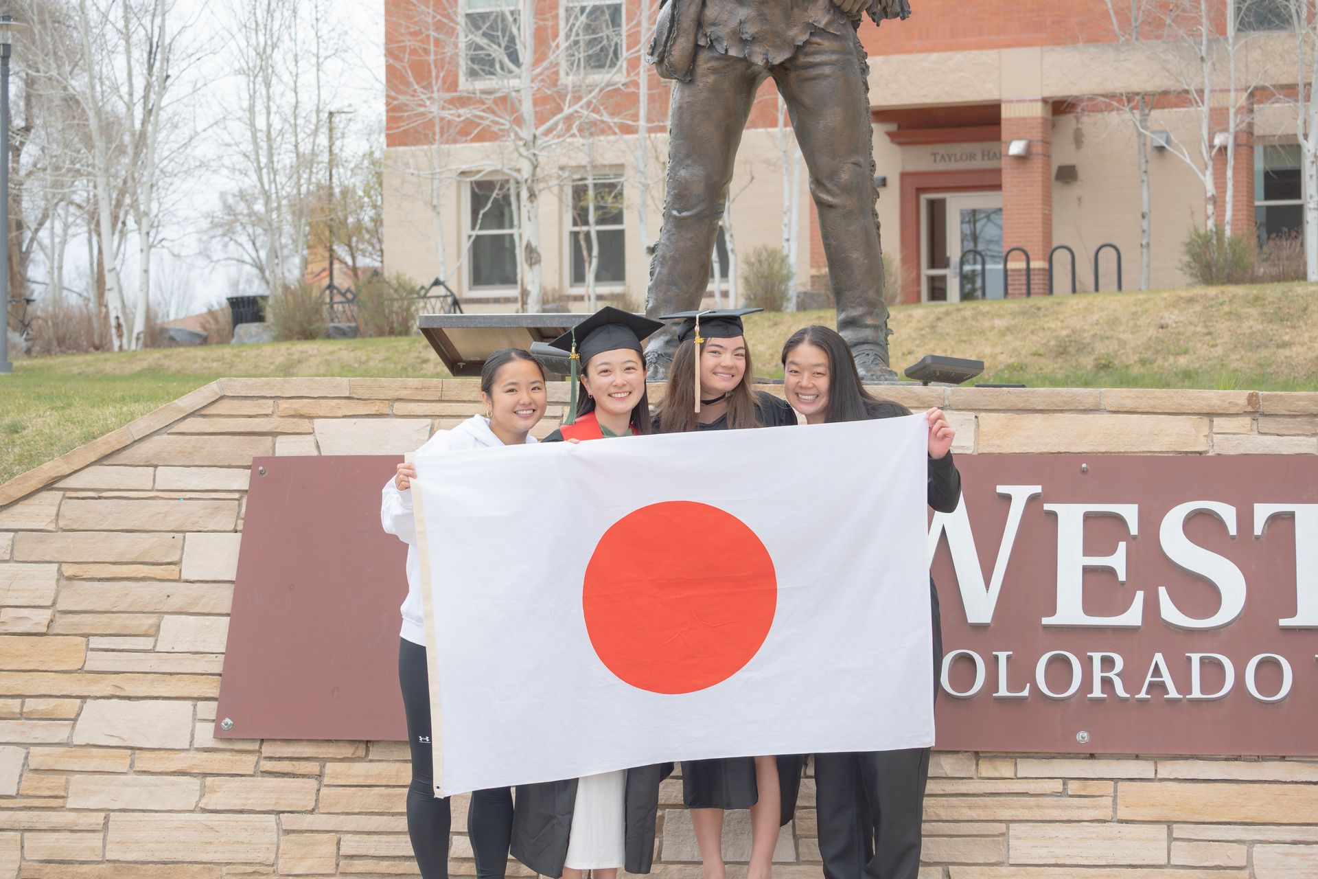 Four graduates in caps and gowns holding a Japanese flag in front of a statue and Western Colorado University sign.