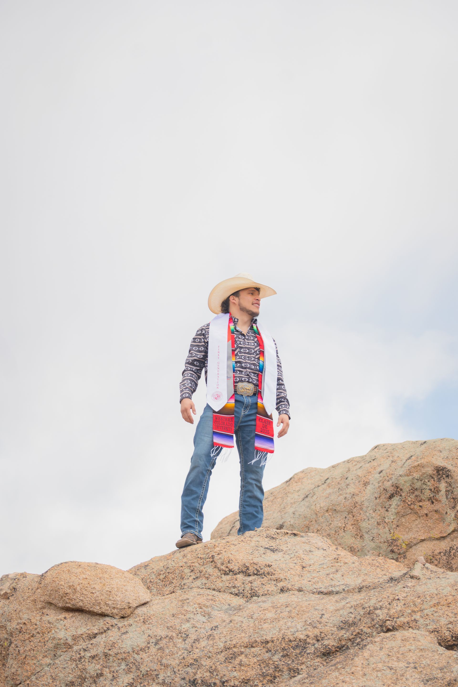 Cowboy in hat stands atop rock, looking to the side. Bright cloudy sky.