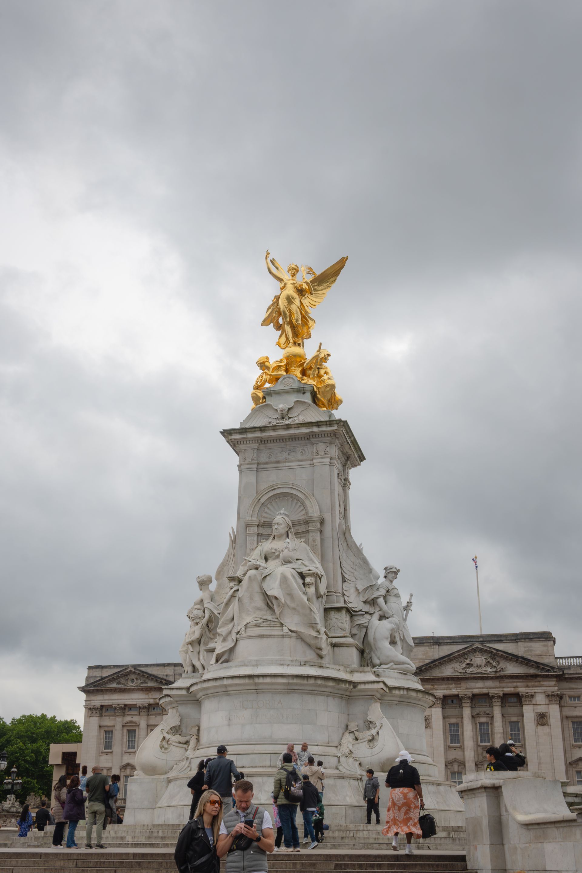 Golden statue of an angel atop marble monument; Buckingham Palace background, cloudy sky.