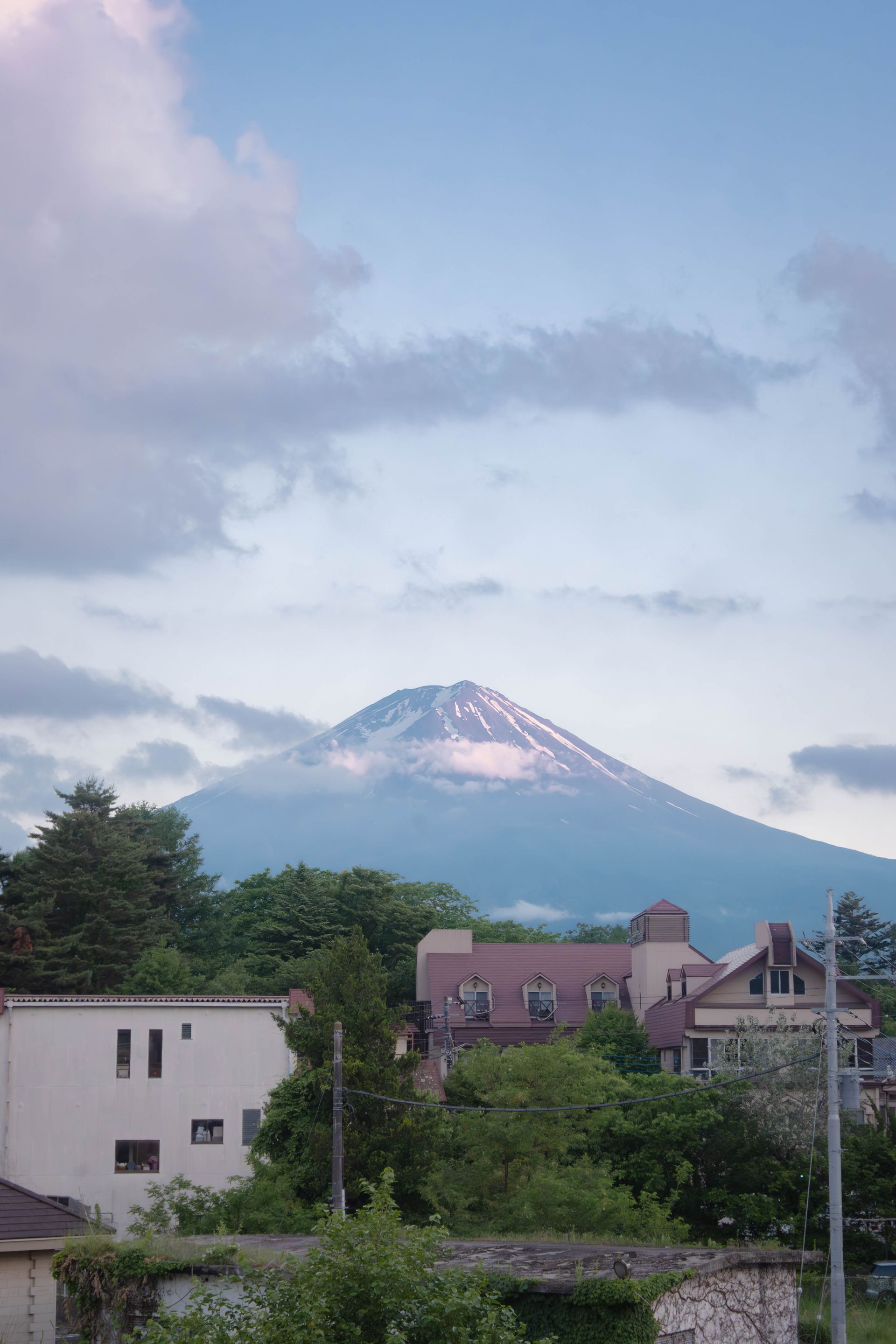 Mt. Fuji rises behind buildings and trees under a cloudy blue sky.