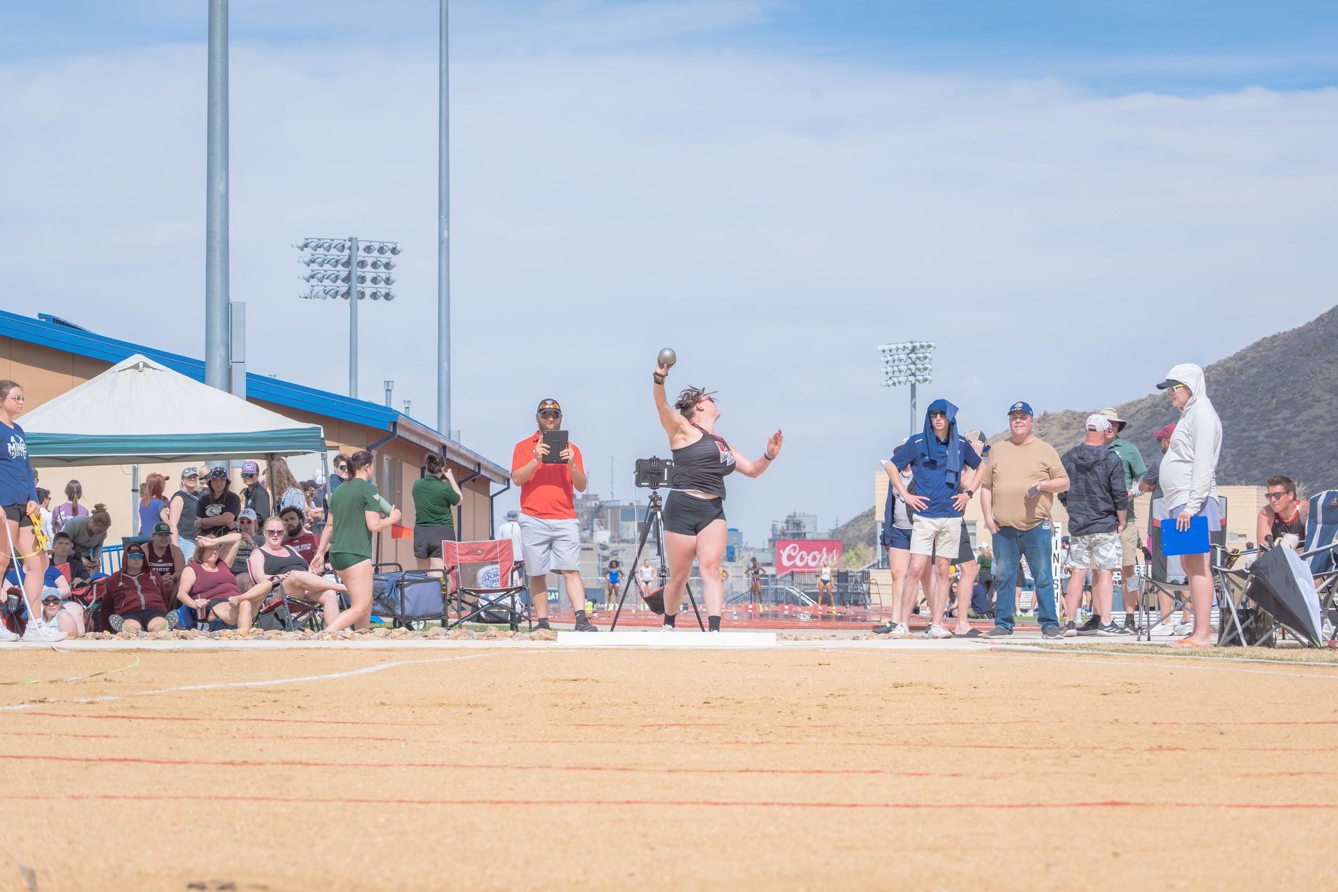 Woman throwing shot put at a track and field event. Outdoors, sunny, with spectators.