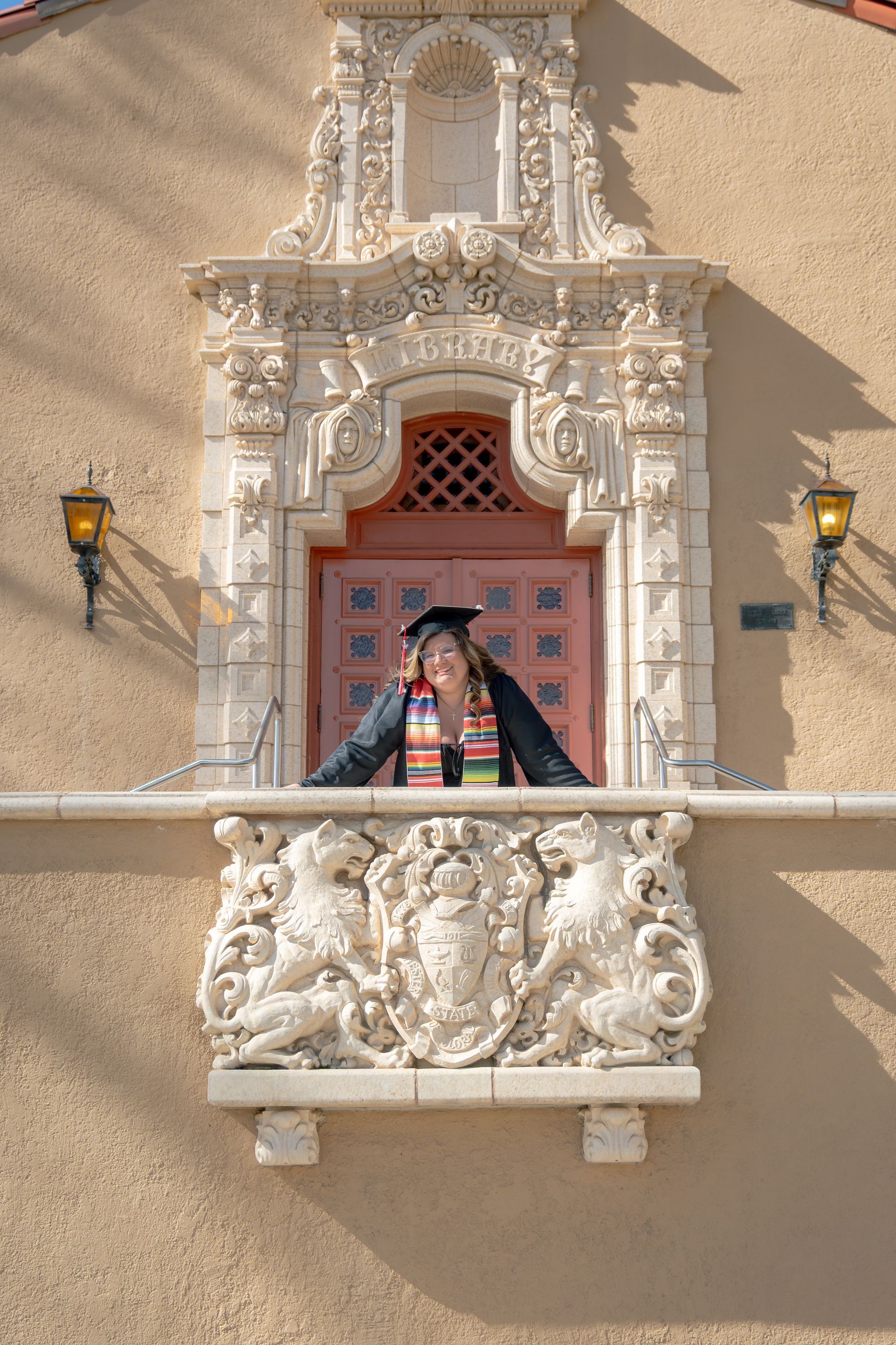 Person in graduation attire in window, arms out, smiling. Ornate stone carvings on beige building.