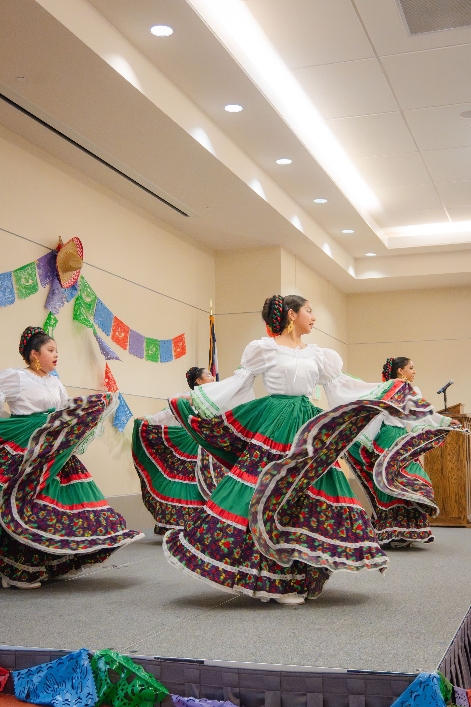 Women in traditional Mexican dresses dance on a stage, skirts swirling.