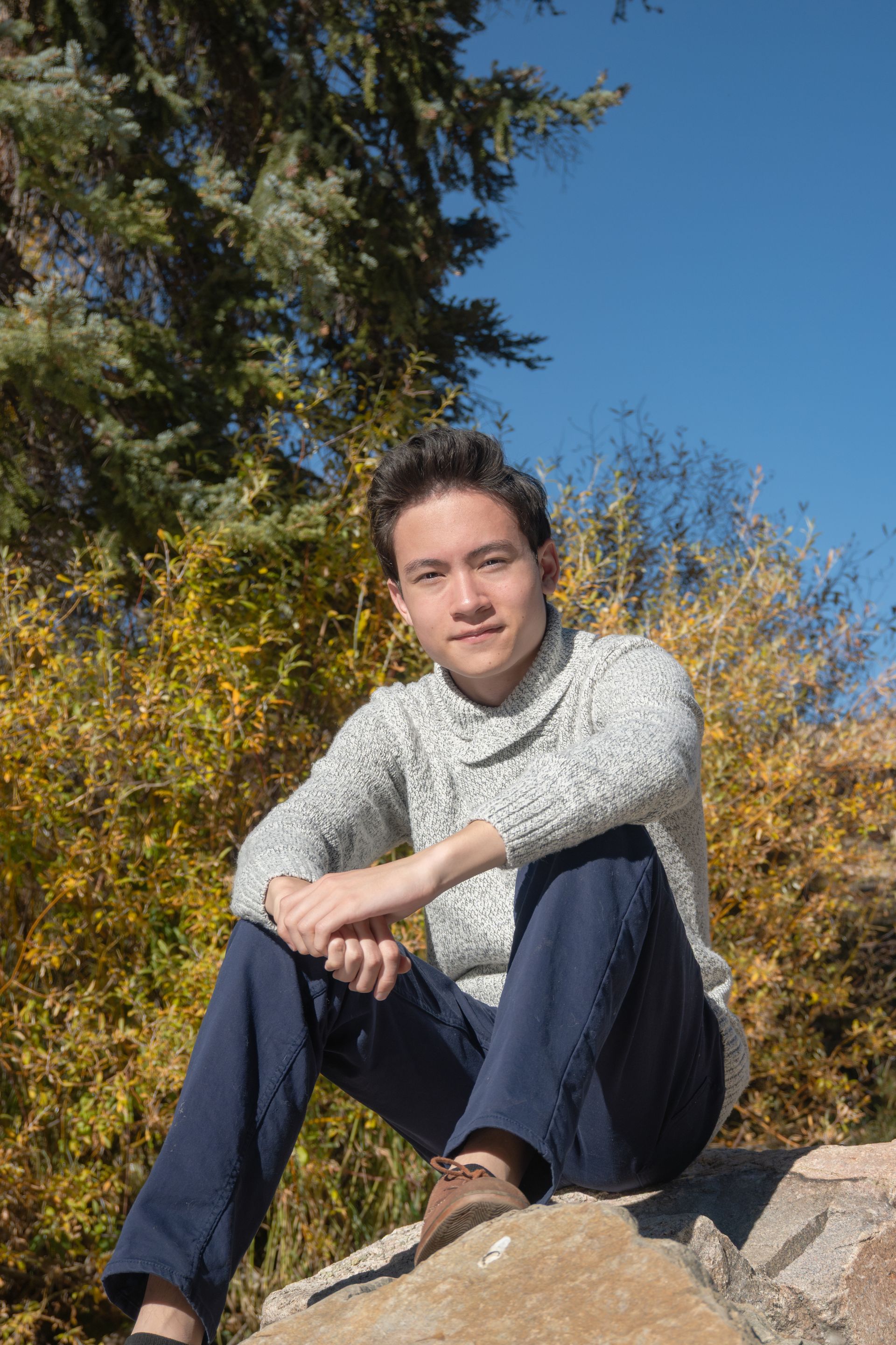 Young man in sweater sits on a rock, arms crossed, looking at the camera, fall foliage background.