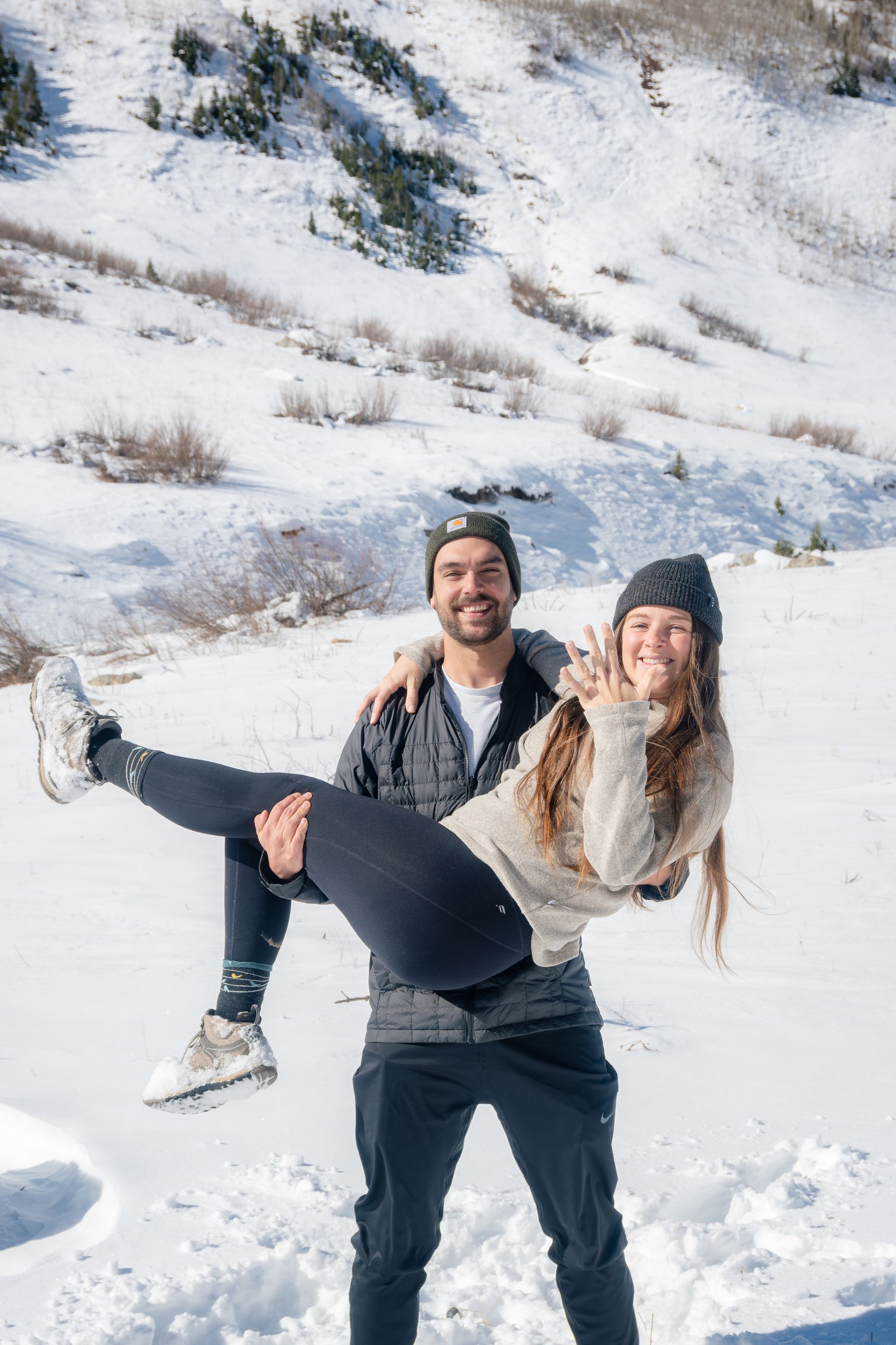 Man holding woman, both smiling in snowy mountains; woman gestures toward camera.