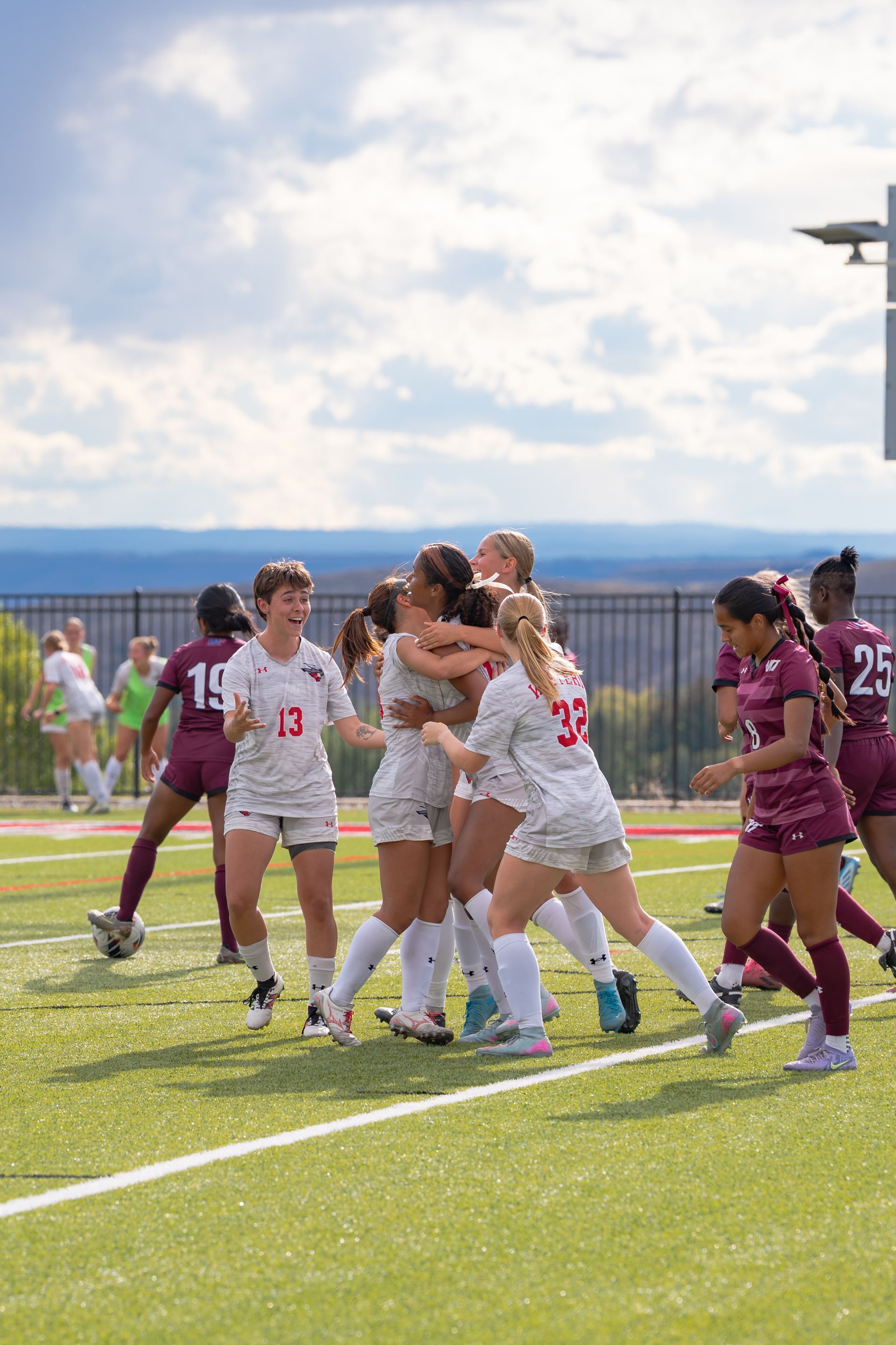 Soccer players in white and red uniforms celebrating a goal on a green field. Other players watch.