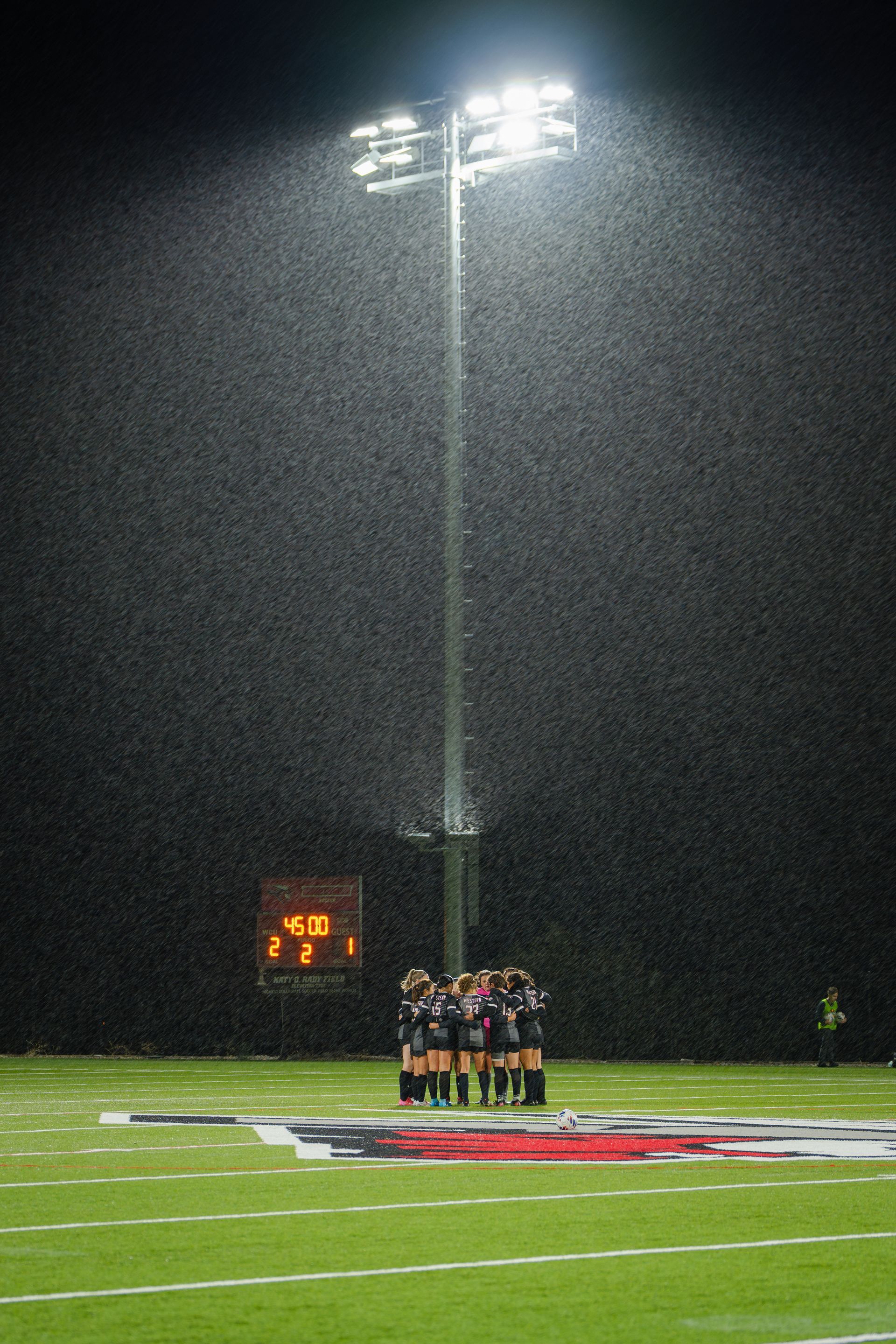 Football team huddles on field during heavy rain under stadium lights at night; scoreboard in background.