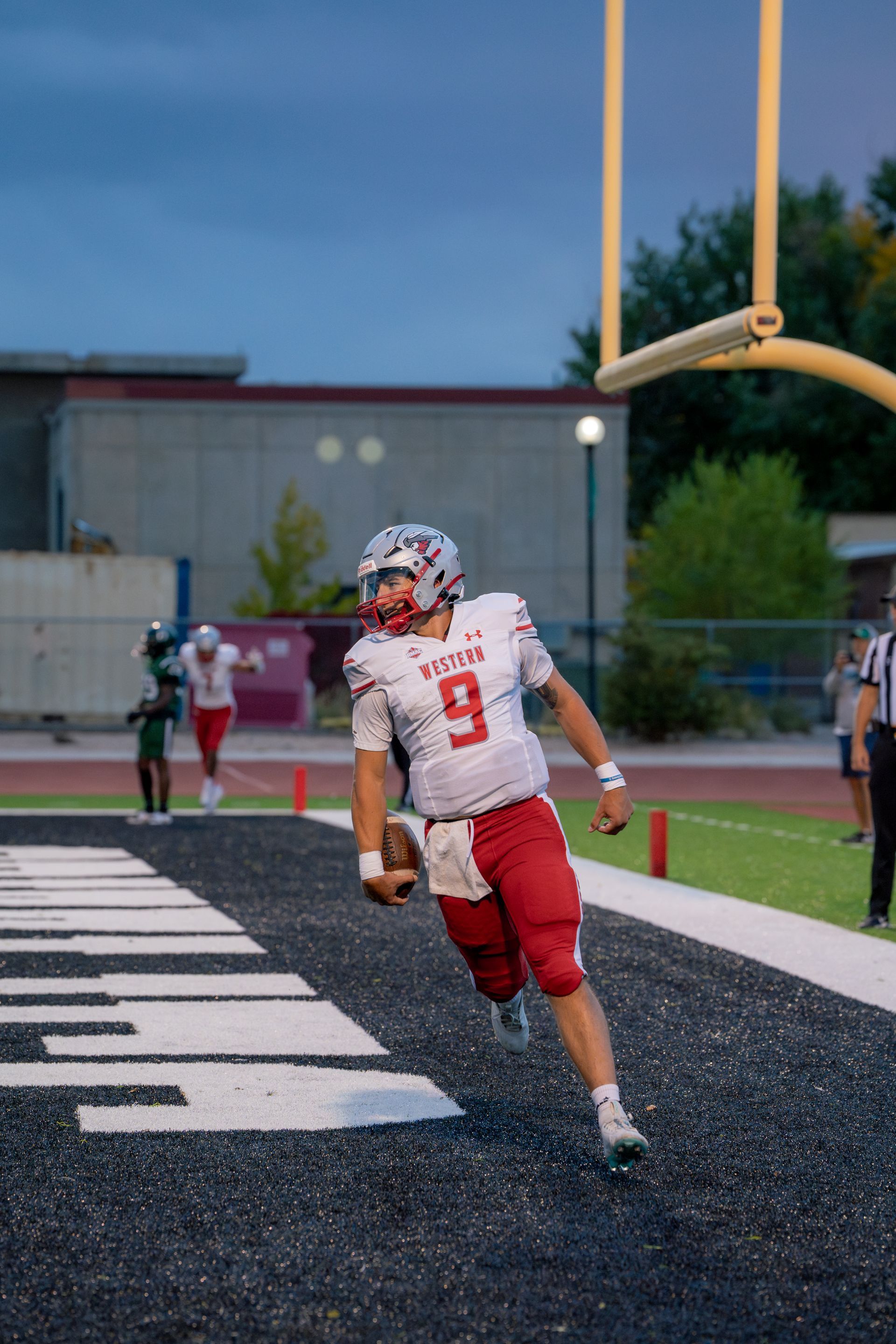 Football player in red uniform, number 9, running into endzone with the ball. Game setting, evening.