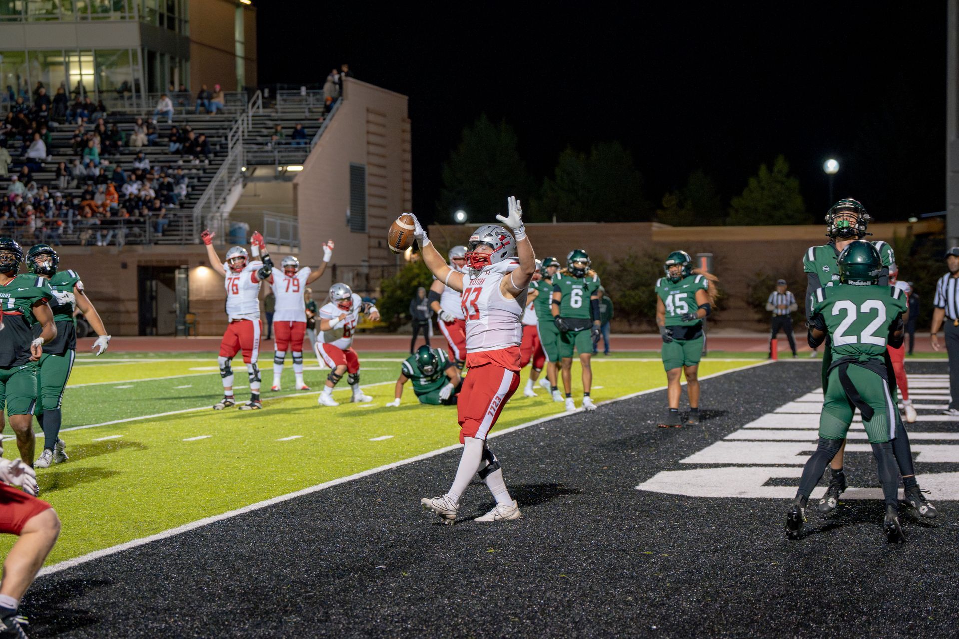 Football player in red celebrates a touchdown in end zone. Teammates cheer. Opponents in green watch.