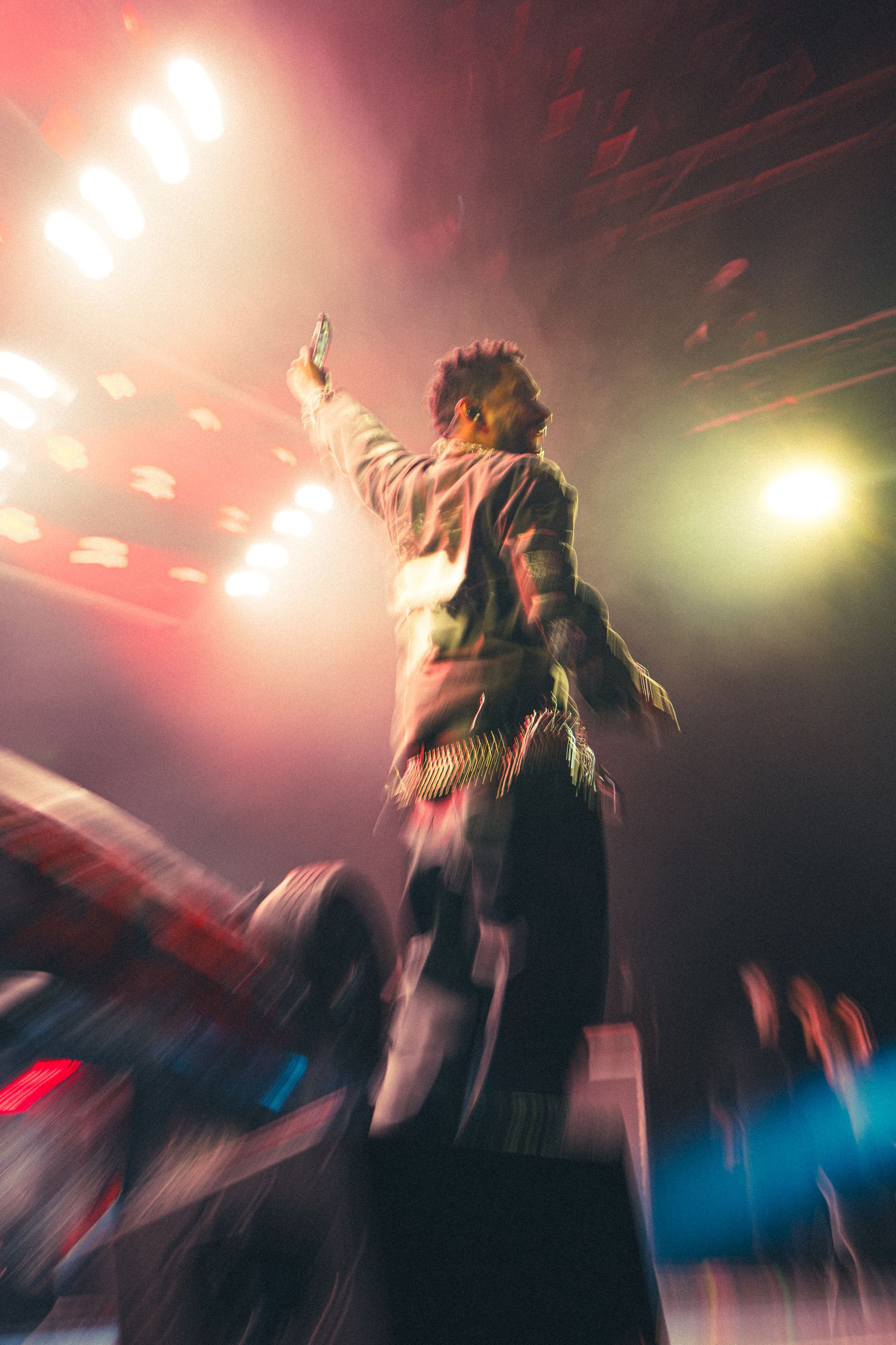 Miguel on stage with arm raised under bright concert lights and colorful smoke