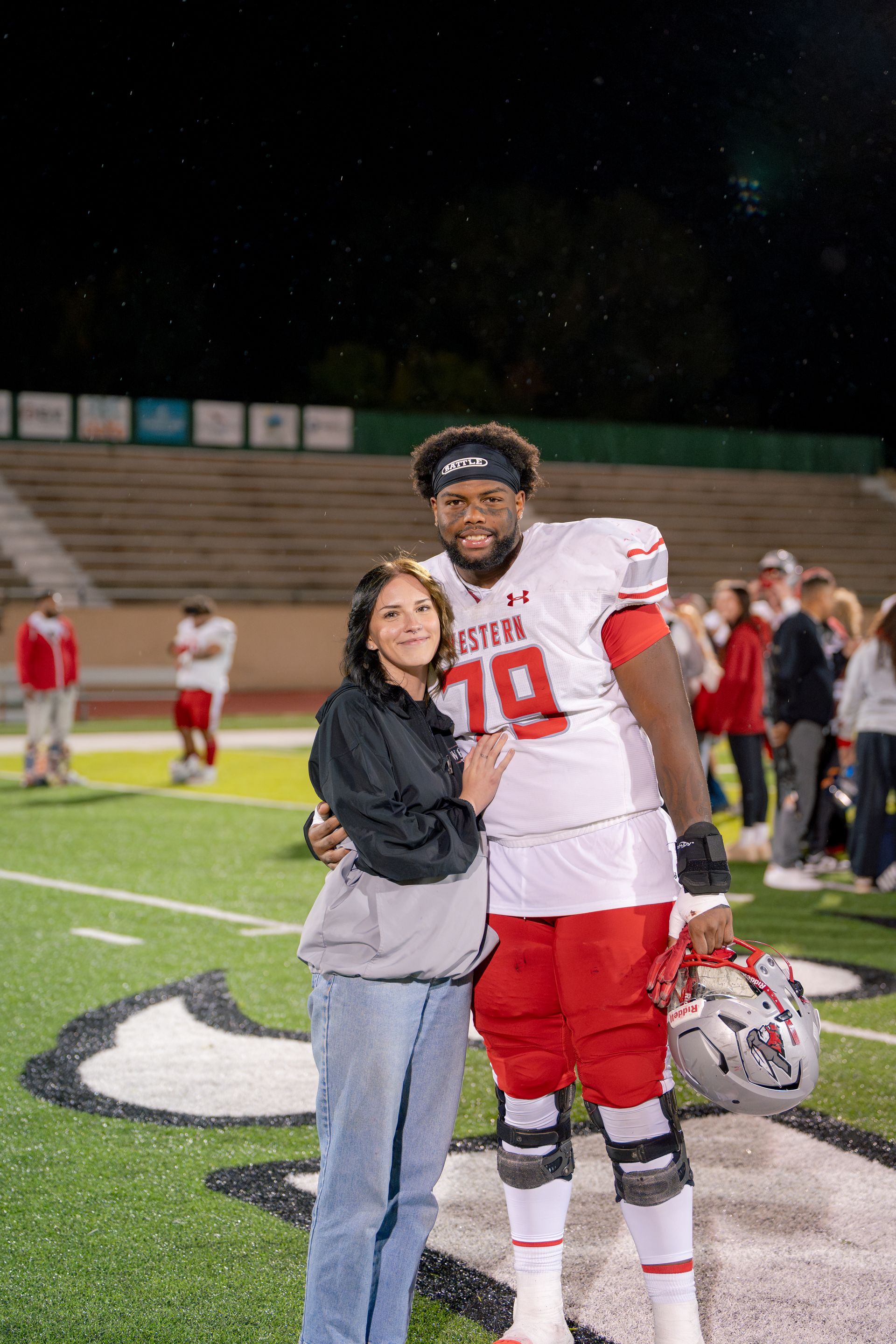Football player in uniform with a woman on the field, smiling. Stadium in background.