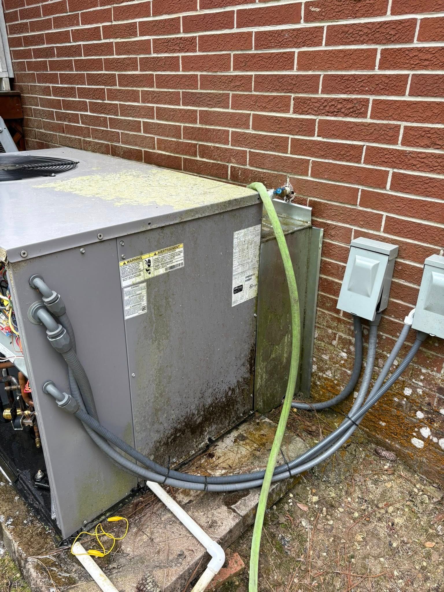 An air conditioner is sitting outside of a brick building with a hose attached to it.