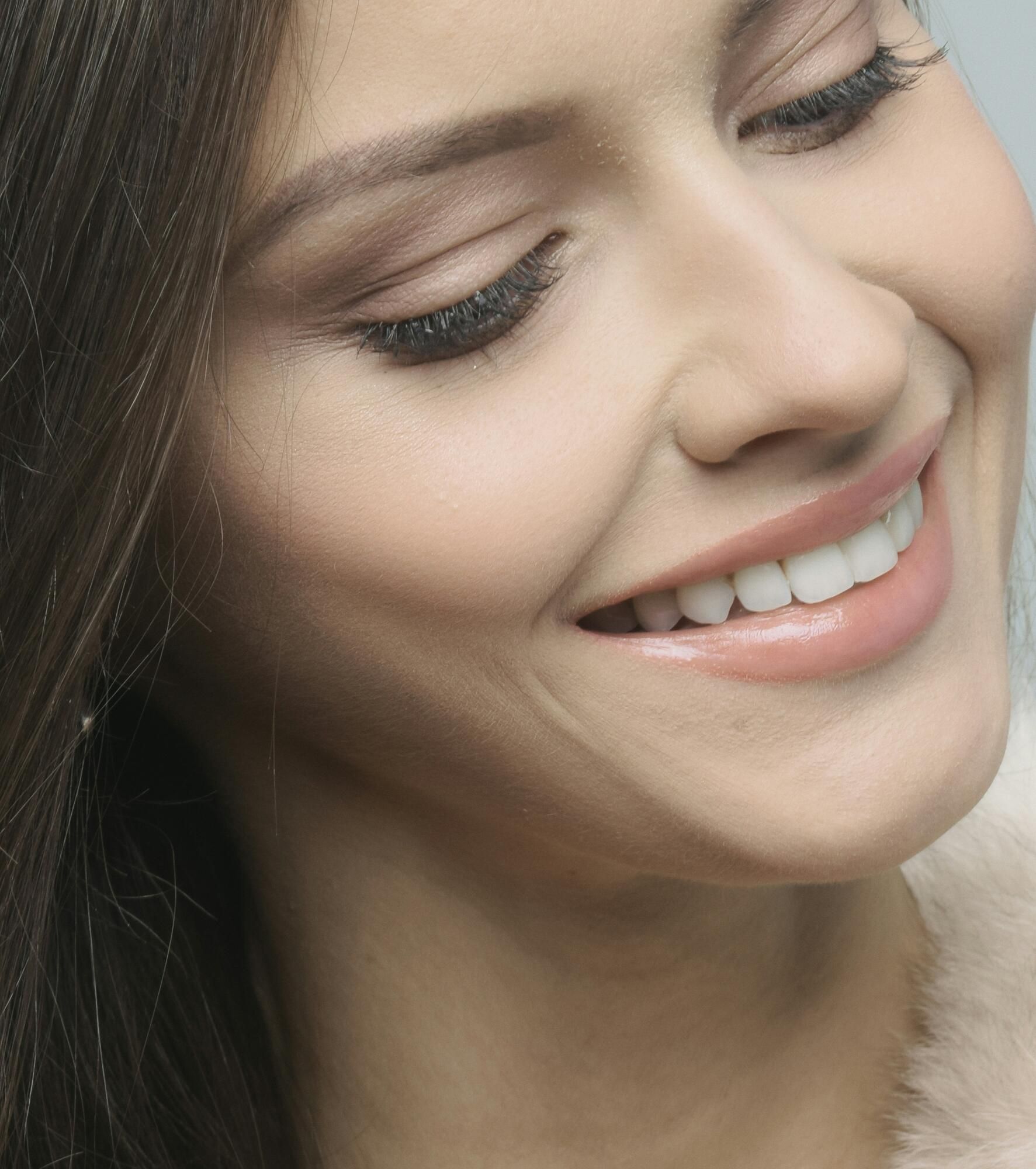 Woman smiling, wearing light makeup, showing teeth, against a soft pink background.