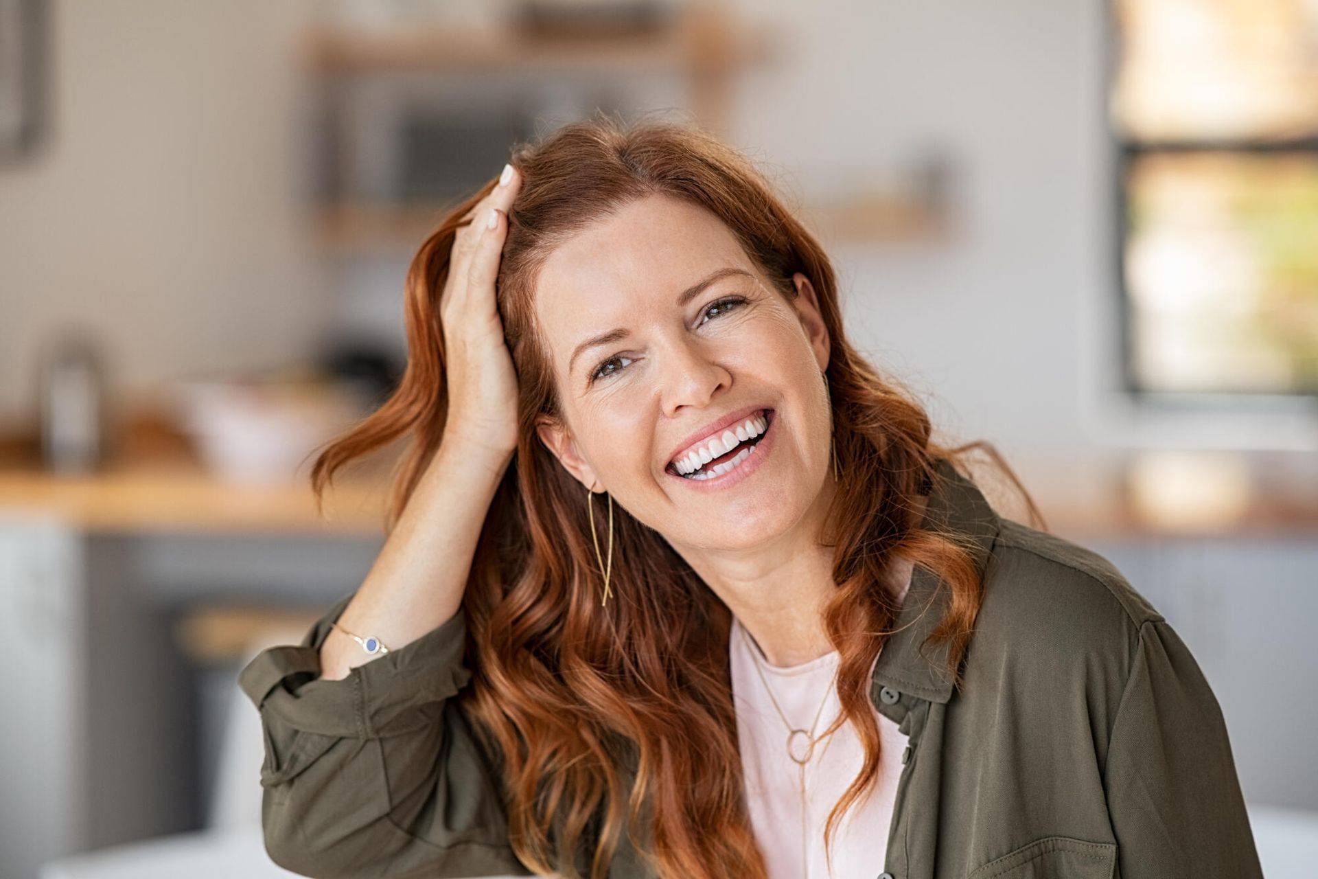 Woman with red hair, smiling, touching her hair, wearing a green jacket, inside a home.