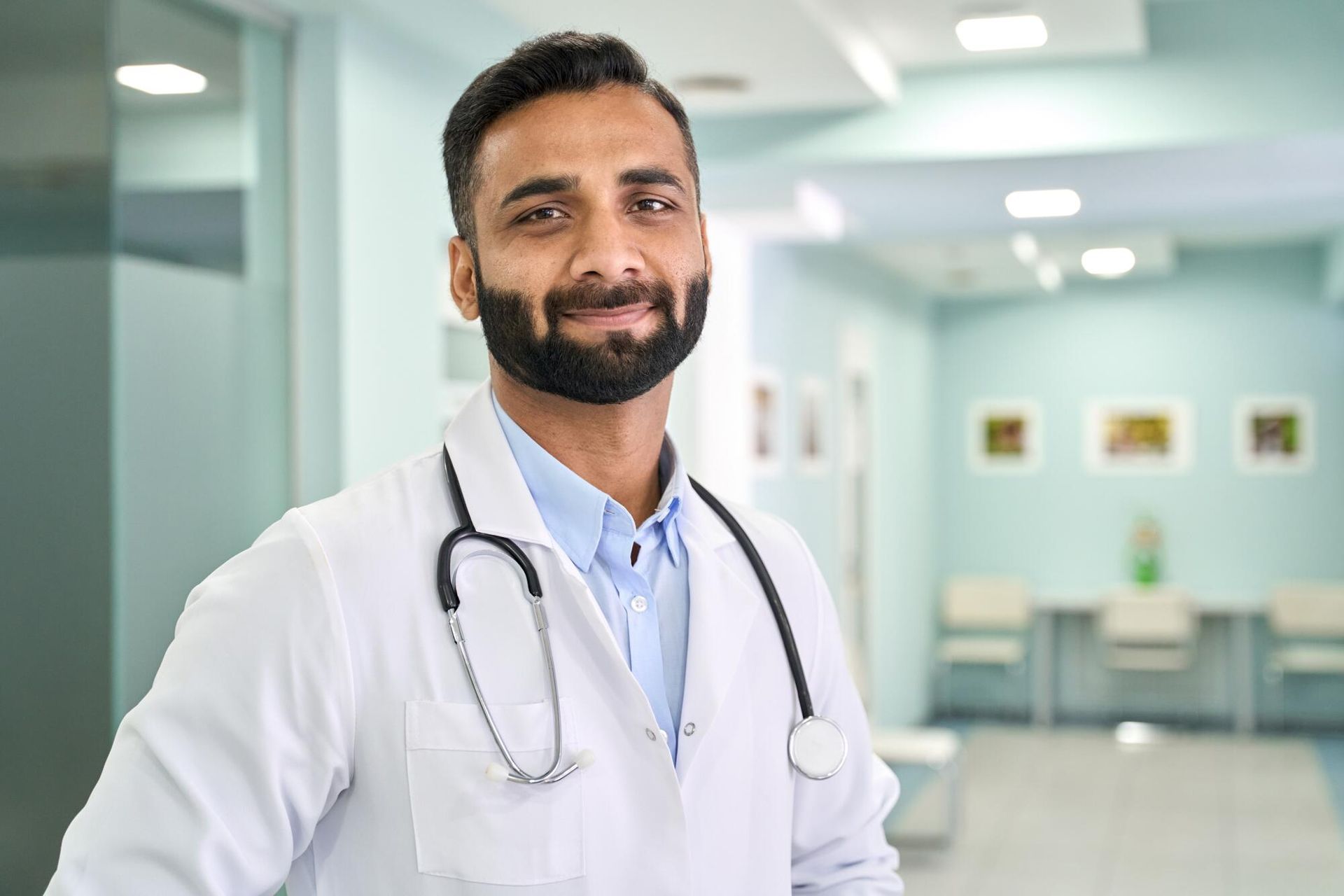 A smiling doctor with a stethoscope around his neck is standing in a hospital hallway.