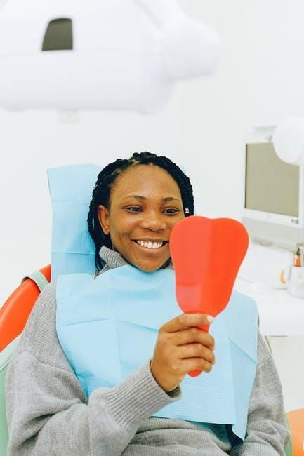 A woman is sitting in a dental chair looking at her teeth in a mirror.