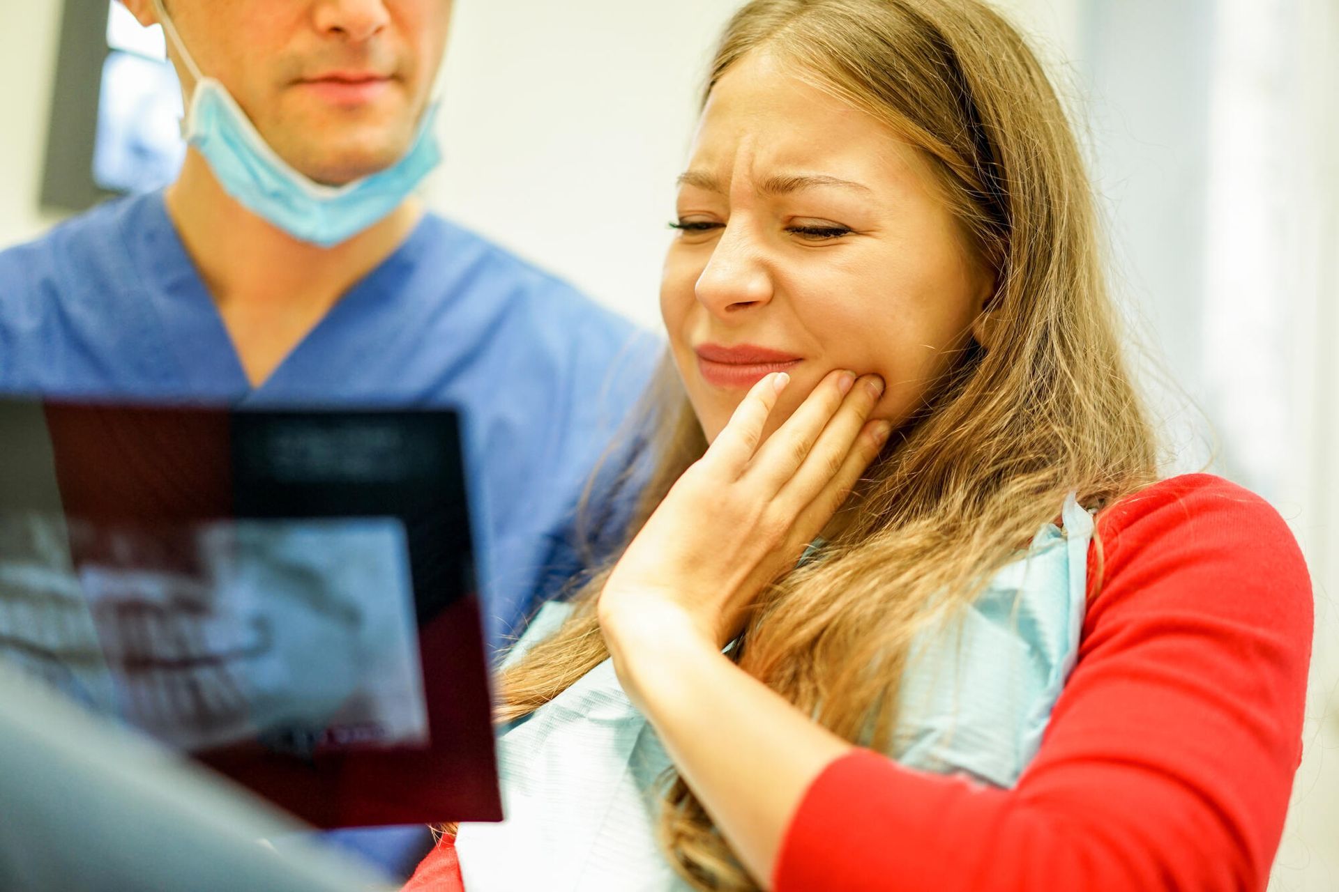 A woman is holding her mouth in pain while a dentist looks at an x-ray of her teeth.