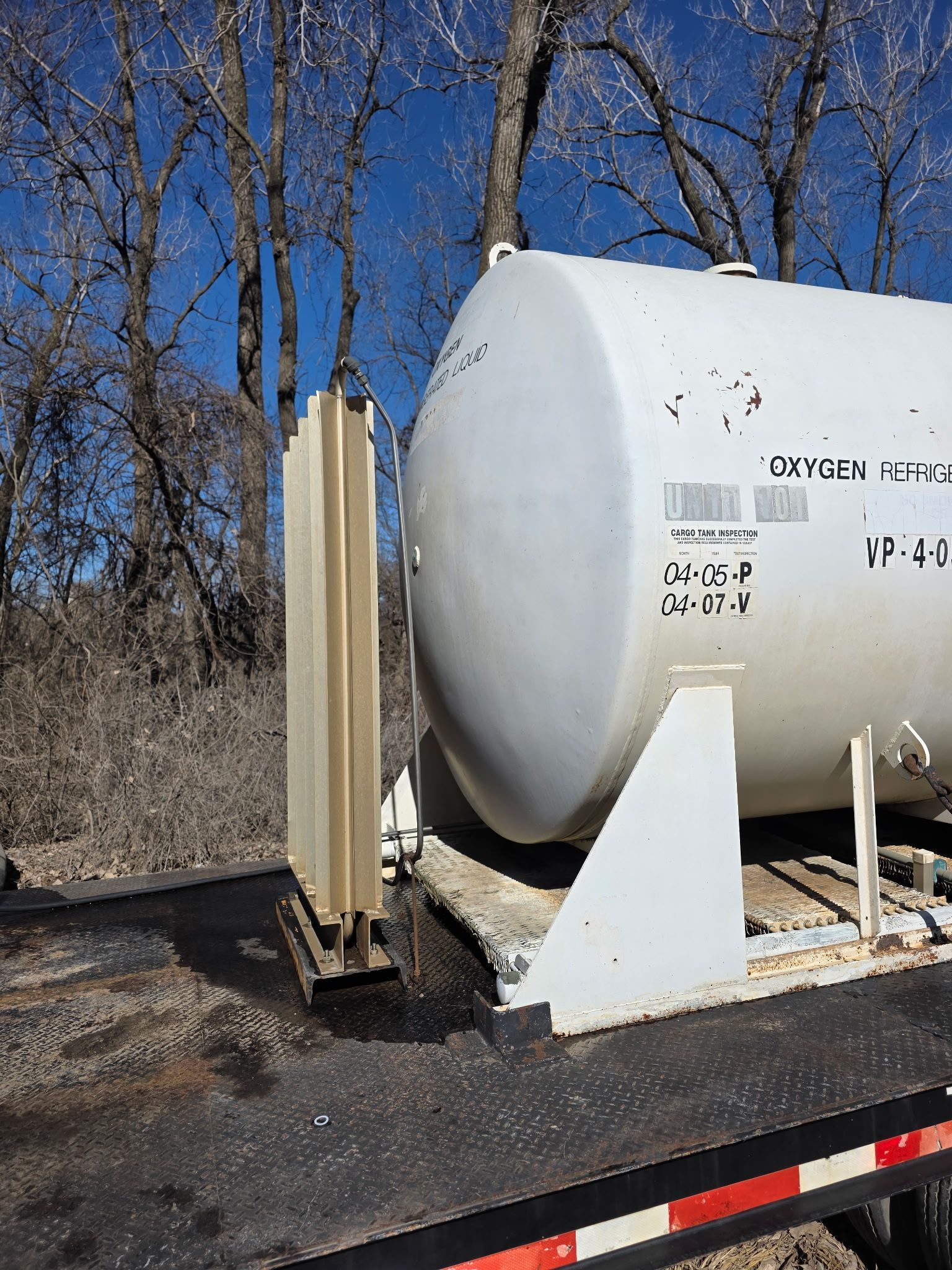 White tank car on a trailer with a tall, metal component on the left side, trees in the background.