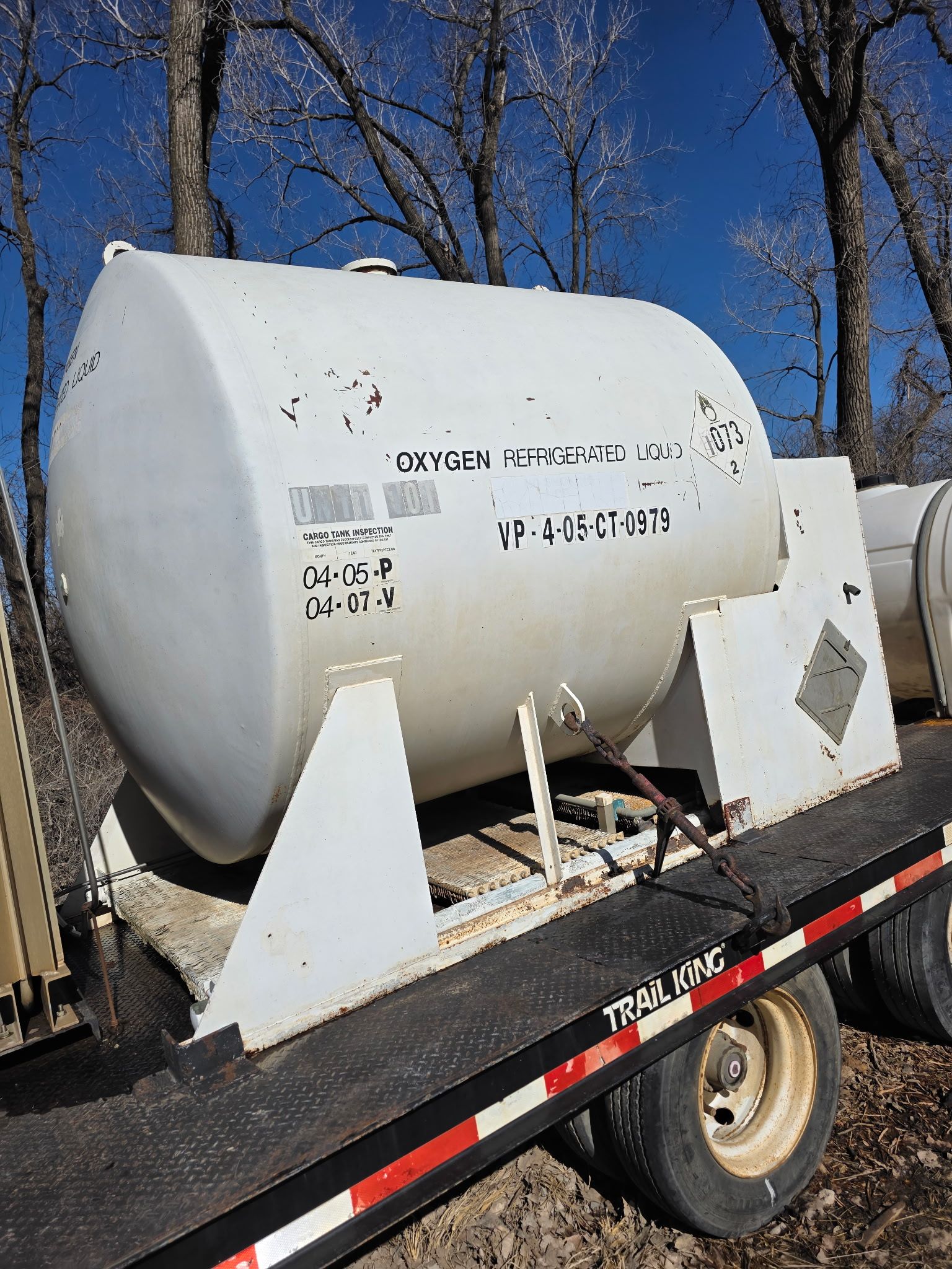 White tank on a trailer with black lettering; outdoors, trees in background, sunny day.