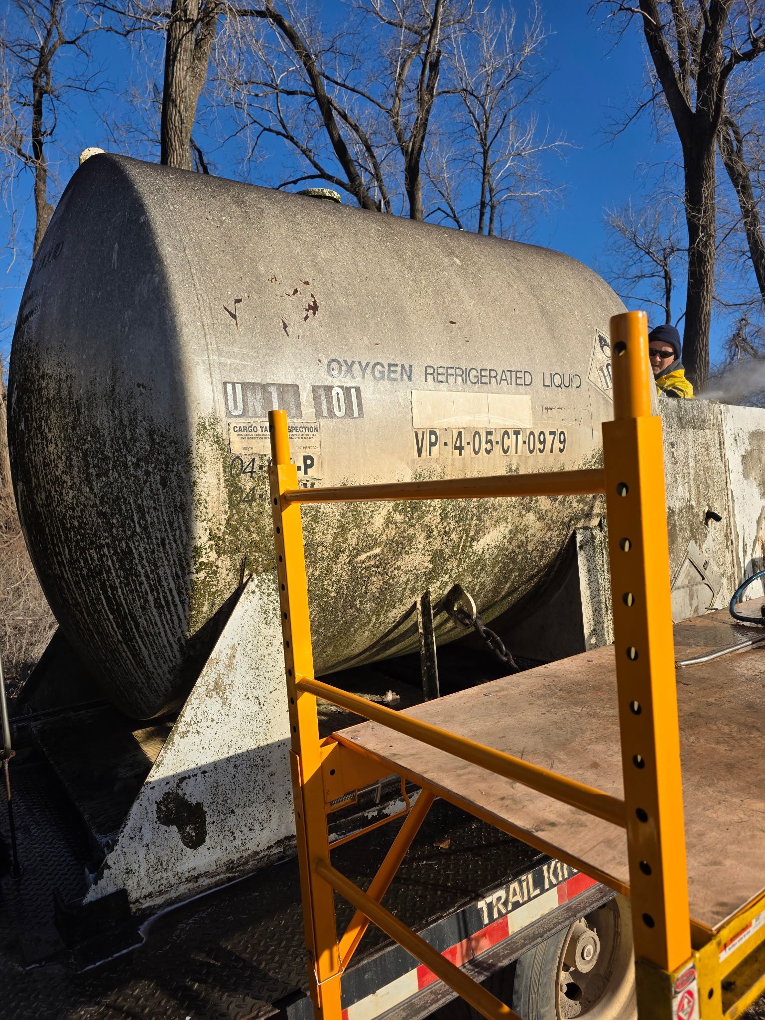 Tanker truck on its side, yellow scaffolding in front. Trees in background.