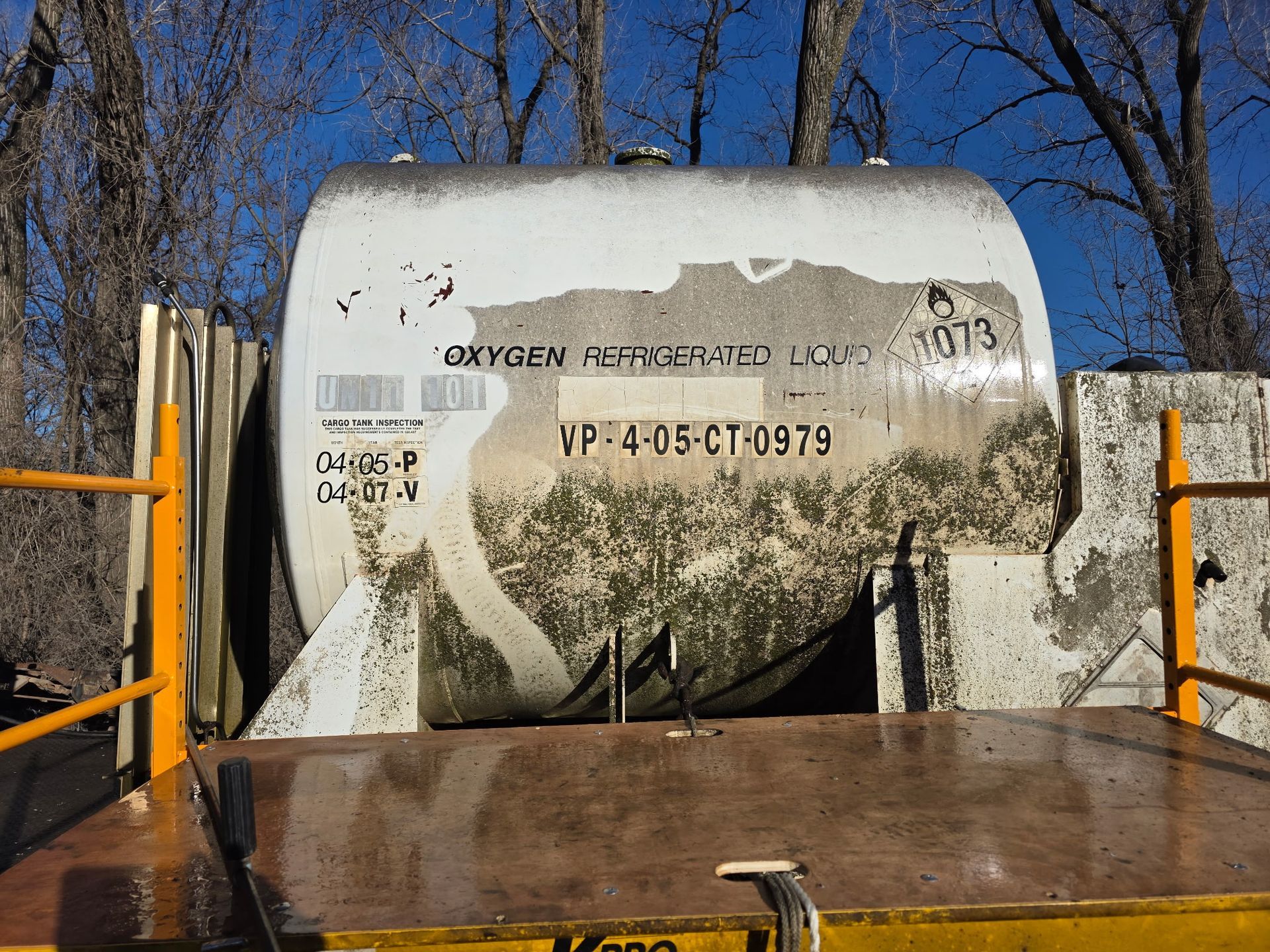 Large white oxygen tank on a yellow flatbed, text partially visible, in front of trees.