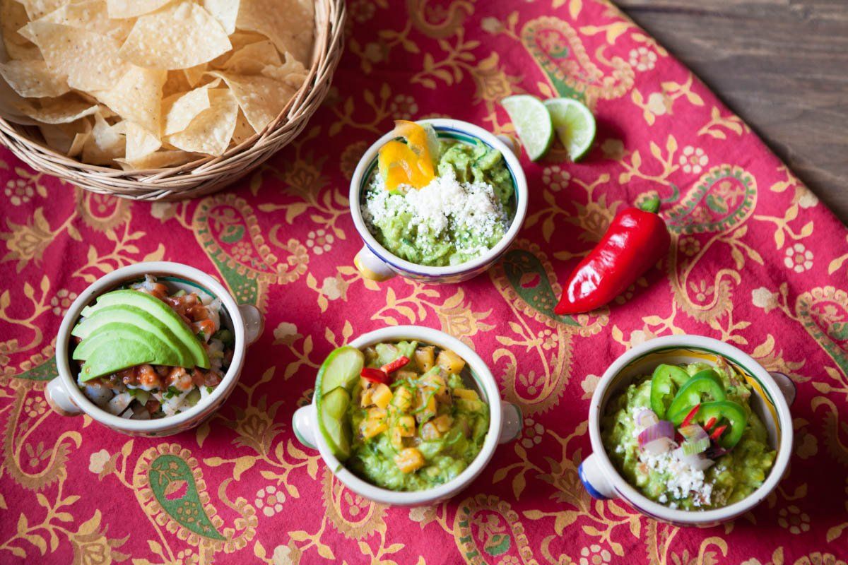 Four bowls of guacamole are sitting on a table next to a basket of tortilla chips.