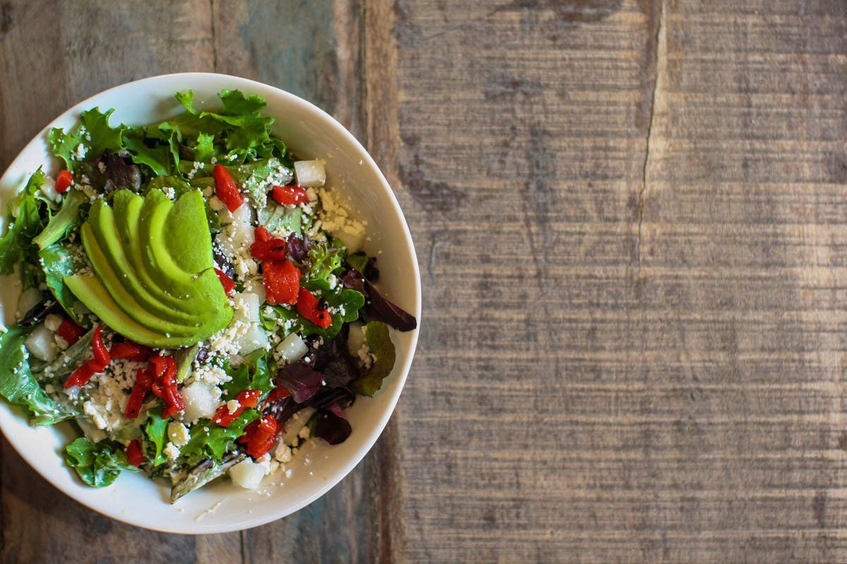 A salad with avocado , lettuce , red peppers and feta cheese on a wooden table.