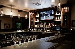 A bar with a television on the wall and martini glasses on the counter.