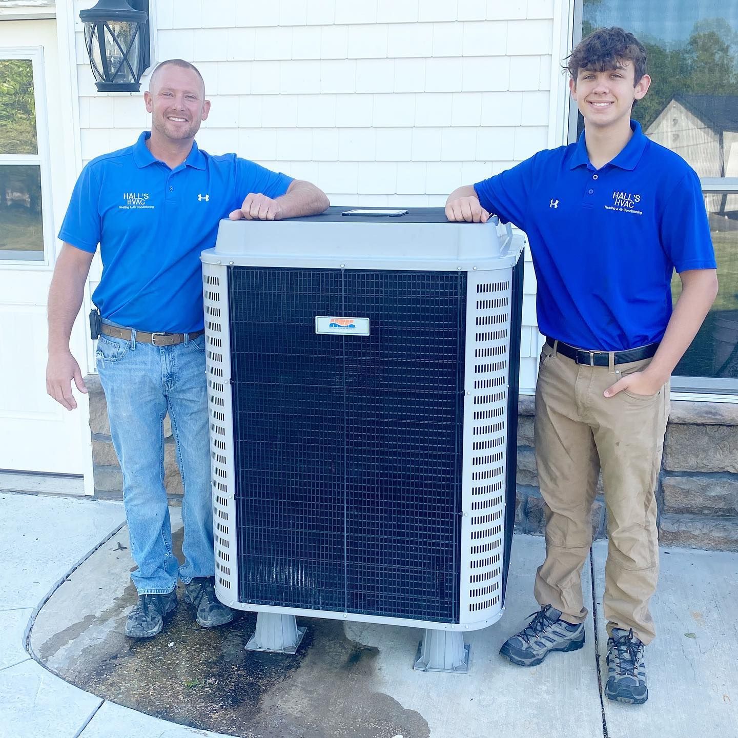 Two men in blue shirts stand with an air conditioning unit outside a building.
