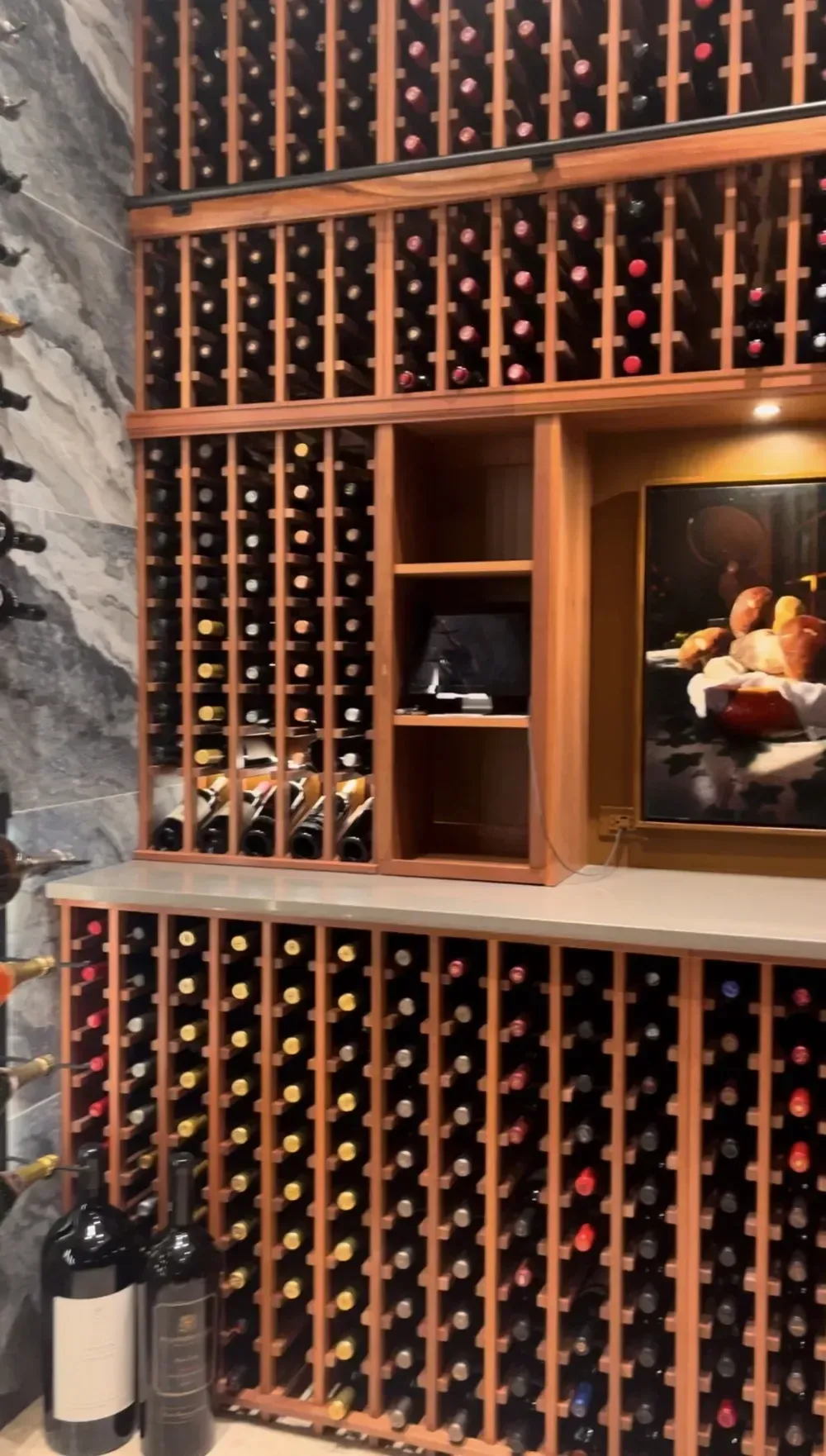 Wooden wine racks filled with bottles, set against a marble wall with a painting in a recessed area.