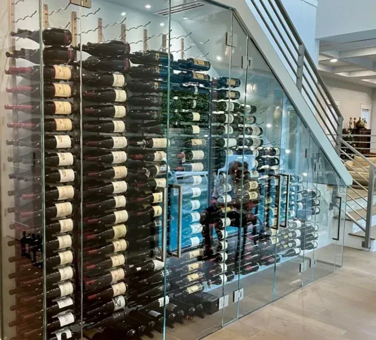 Glass-enclosed wine cellar built under a staircase, displaying rows of wine bottles in racks.