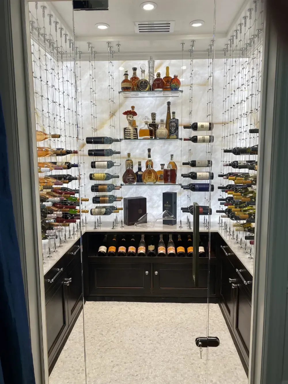 Wine cellar with bottles displayed on shelves and in cabinets. Marble walls and flooring. Glass door.
