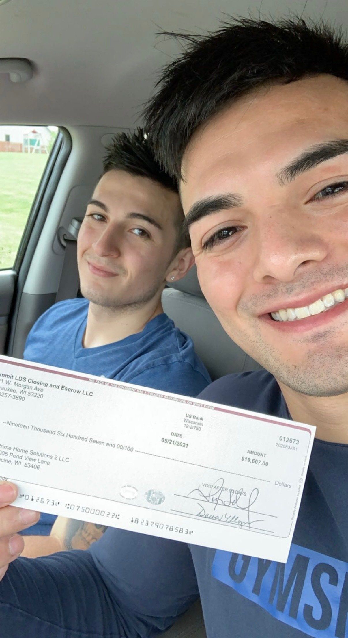 Two men are sitting in a car holding a check.
