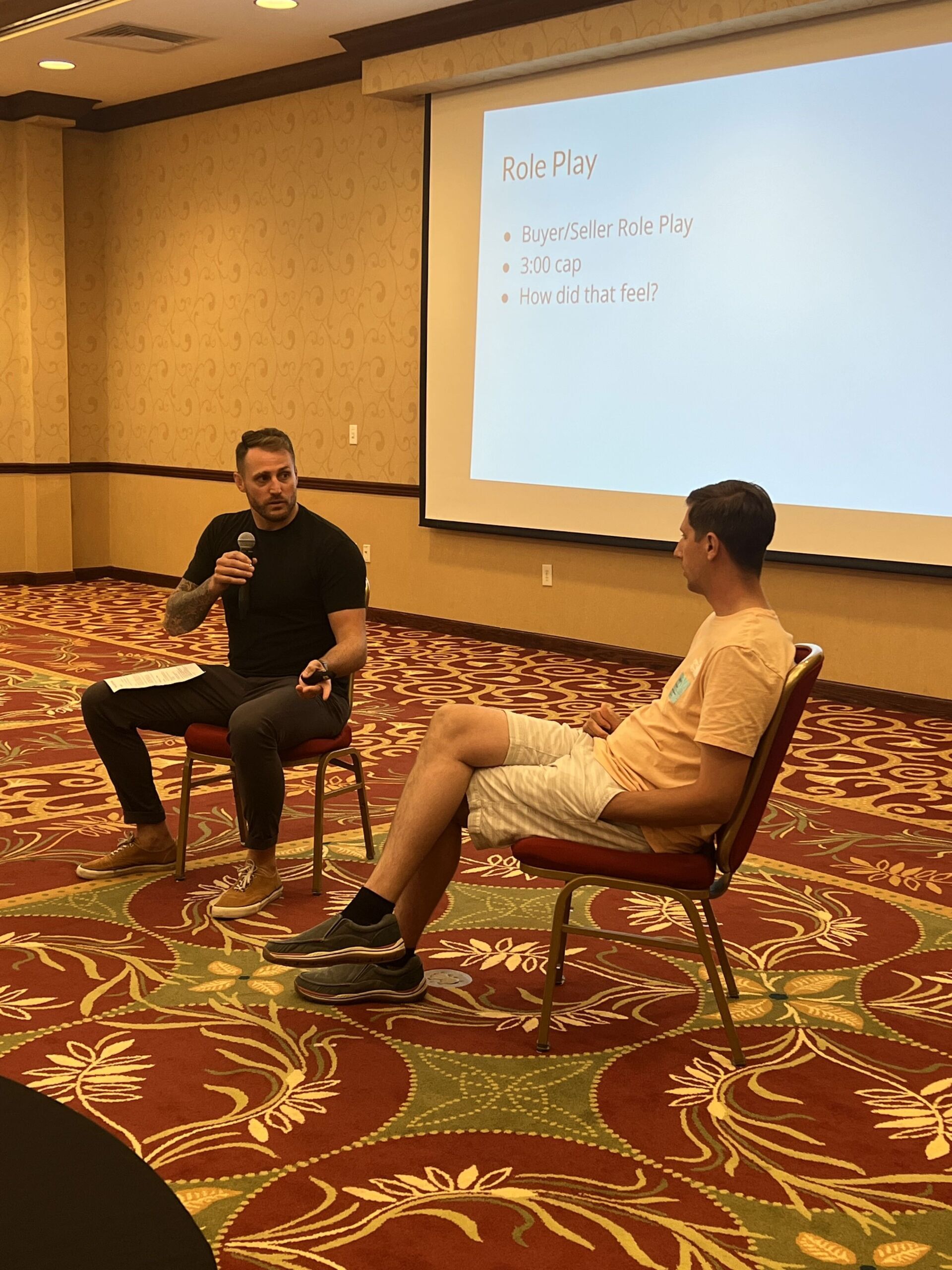 Two men are sitting in chairs talking in front of a projector screen.