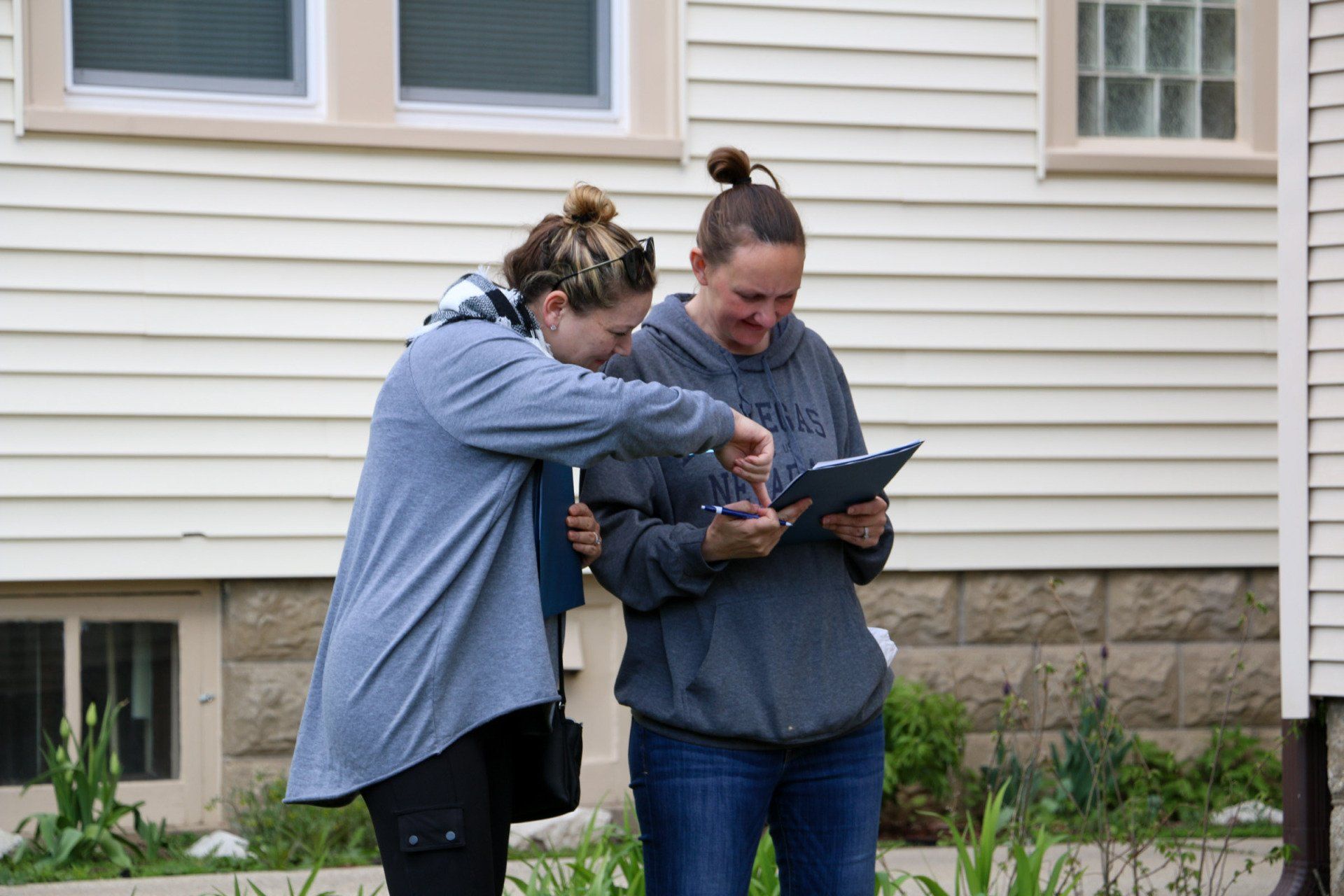 Two women are standing in front of a house looking at a tablet.