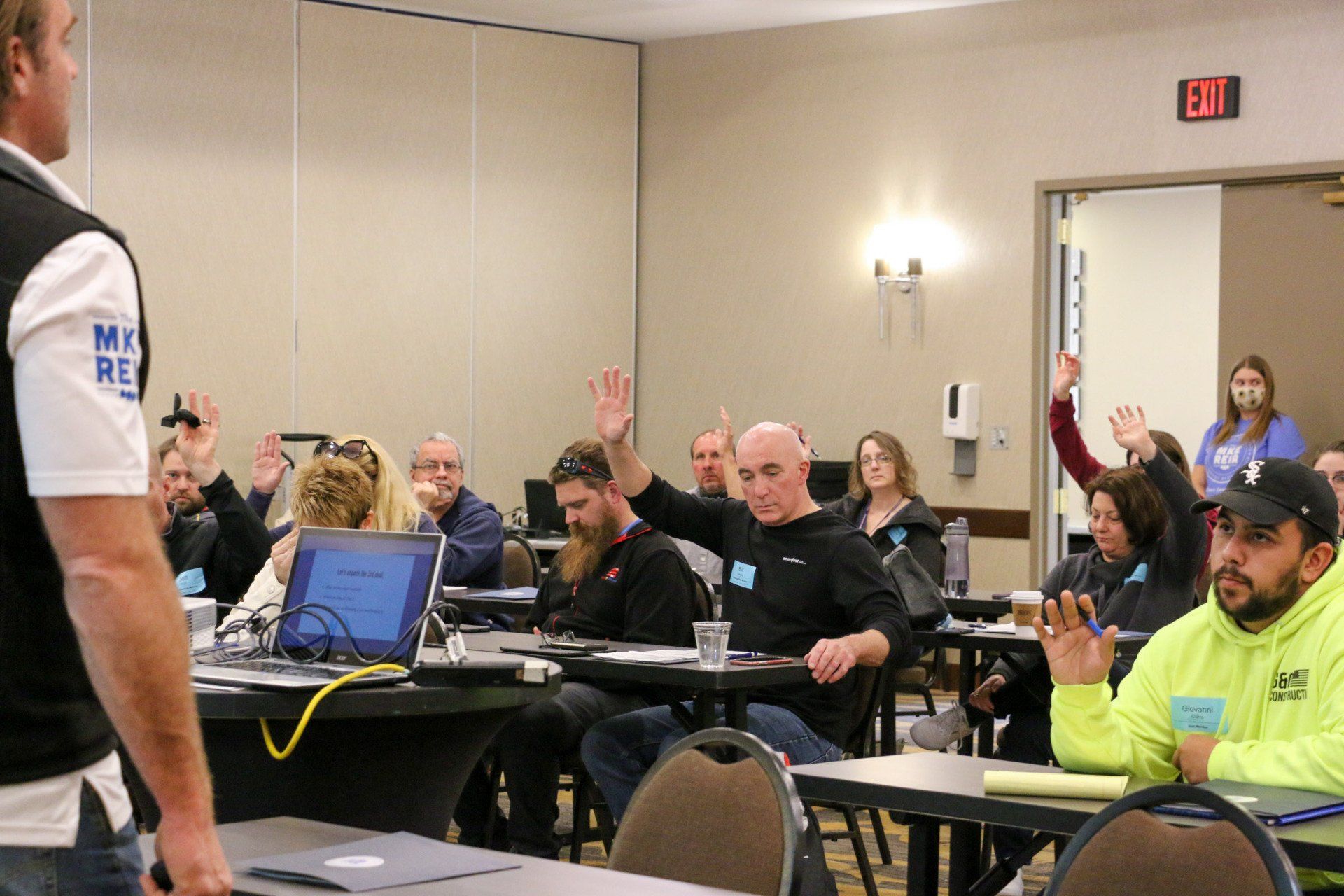A man is standing in front of a group of people sitting at tables raising their hands.
