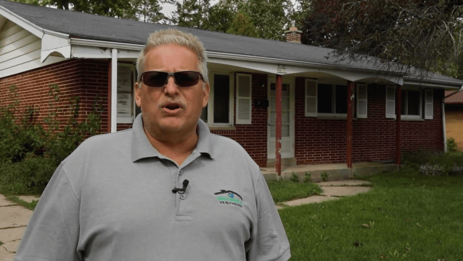 A man wearing sunglasses is standing in front of a brick house.