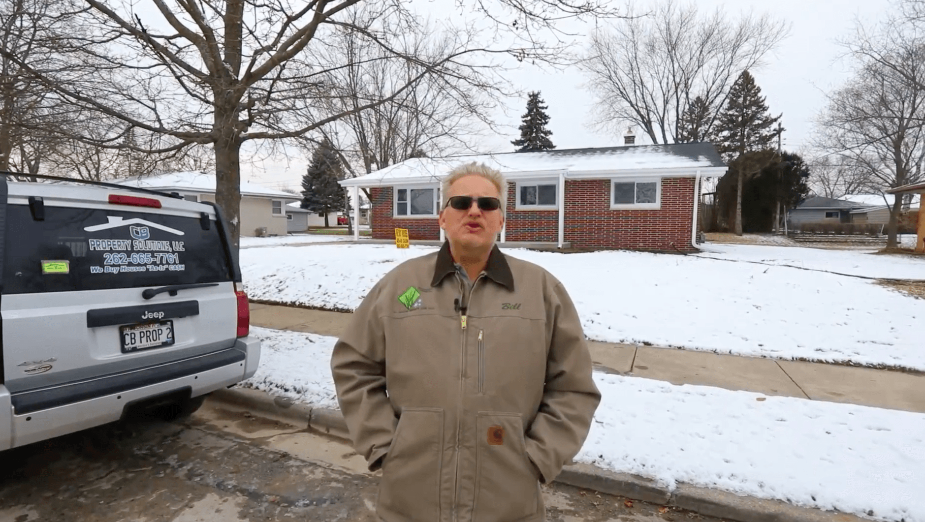 A man wearing sunglasses is standing in front of a snowy house.