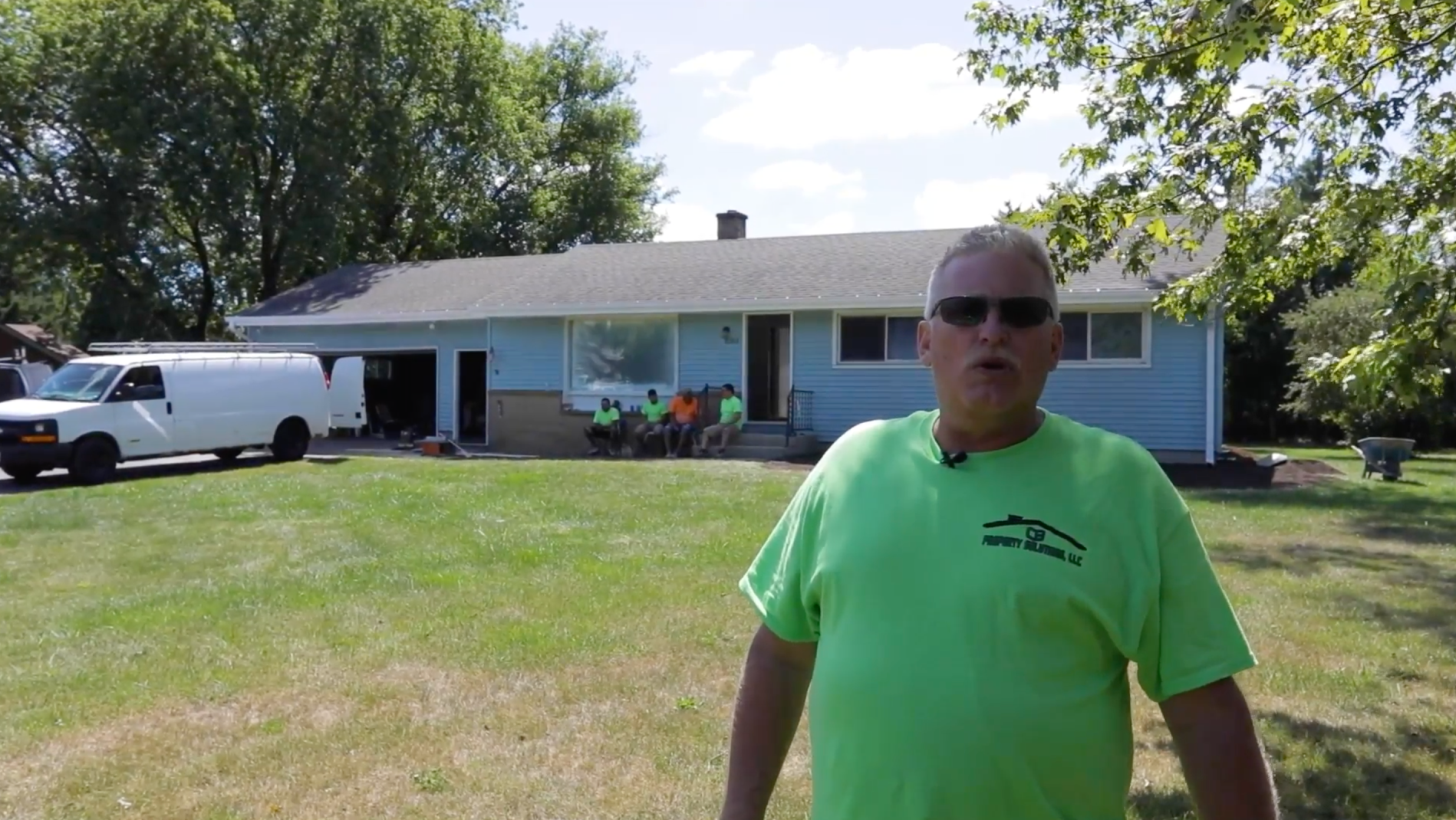 A man in a green shirt is standing in front of a blue house.