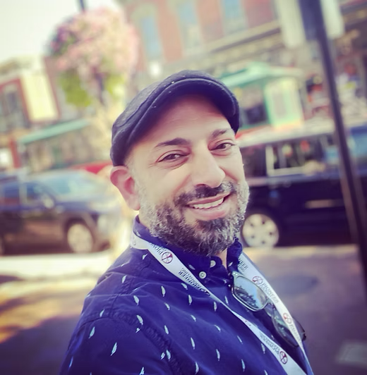 Man with a beard smiles outdoors, wearing a blue shirt and cap; buildings and cars in background.