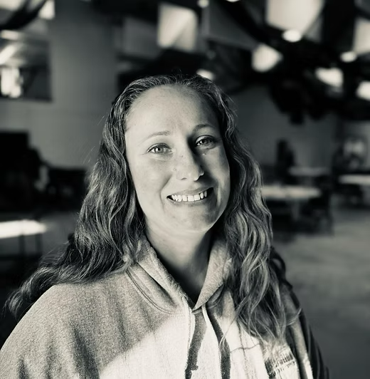 Woman with long hair smiles, wearing a hooded sweatshirt in a blurry indoor setting.