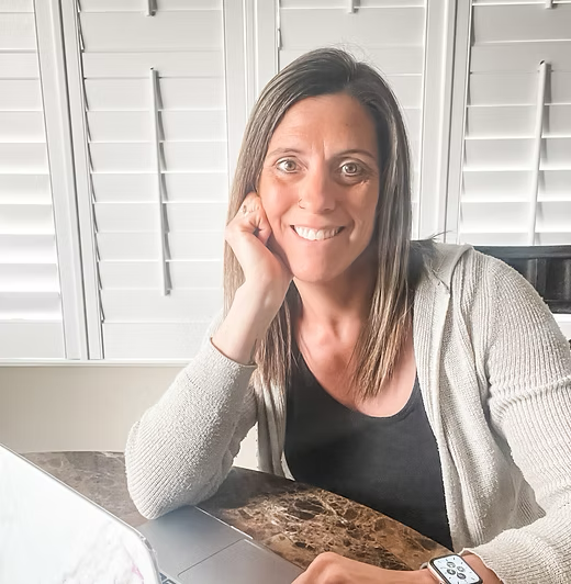 Woman smiling, leaning on her hand, working at a laptop near a window with white blinds.